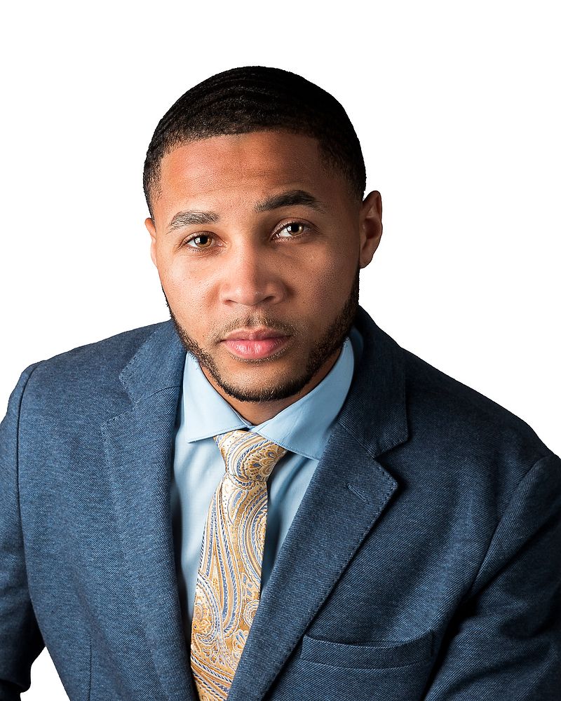Professional headshot of a person in a navy suit jacket, light blue dress shirt and patterned gold tie against white background.