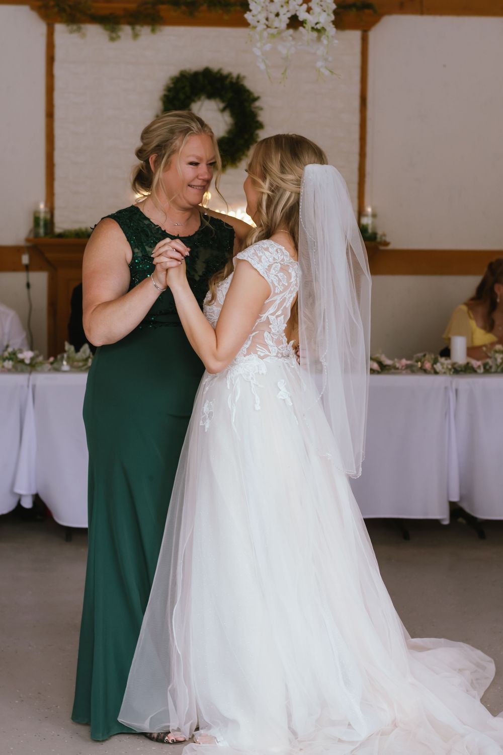Two figures share a joyful moment while dancing at a wedding reception, with a wreath and white tables in the background.