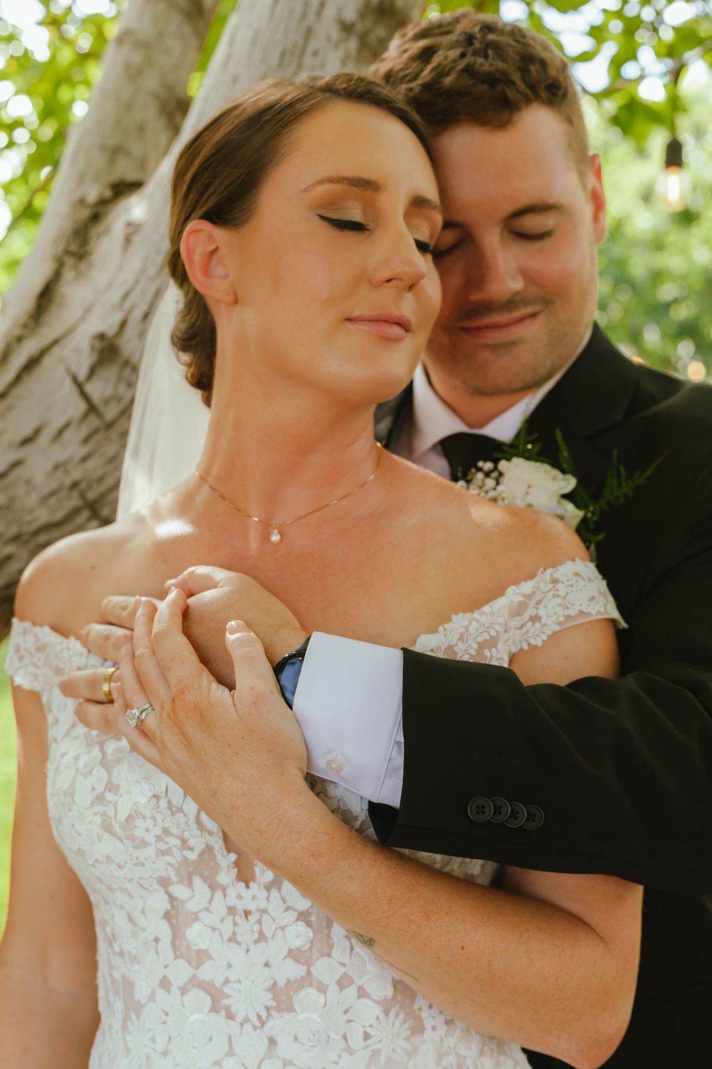 A couple shares an intimate moment on their wedding day, embracing under a tree in natural sunlight.