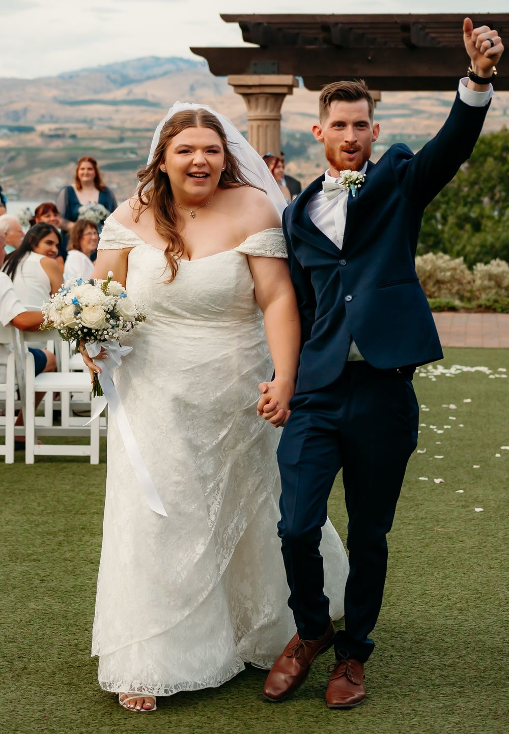 A newlywed couple celebrates joyfully at their mountain venue wedding with blue skies and decorative pillars behind them.