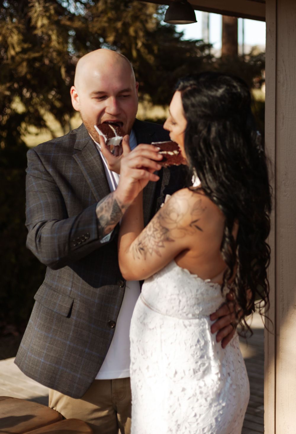 A newlywed couple sharing a piece of cake during their wedding celebration outdoors.