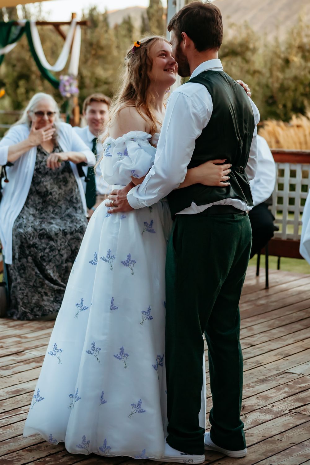 Couple shares a dance moment on wooden deck wearing formal green and white attire at outdoor celebration.