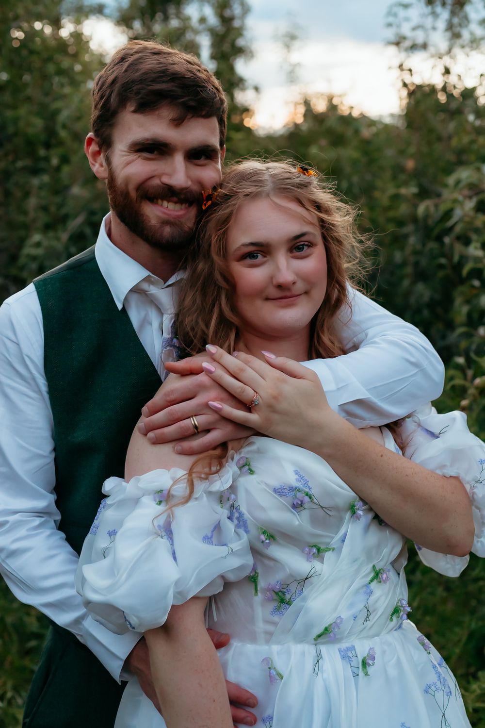 A romantic couple embraces outdoors in formal attire featuring a green vest and floral white dress at sunset.