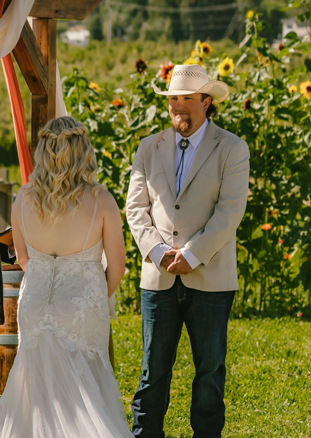 A rustic outdoor wedding ceremony with a backdrop of bright sunflowers and greenery on a sunny summer day.