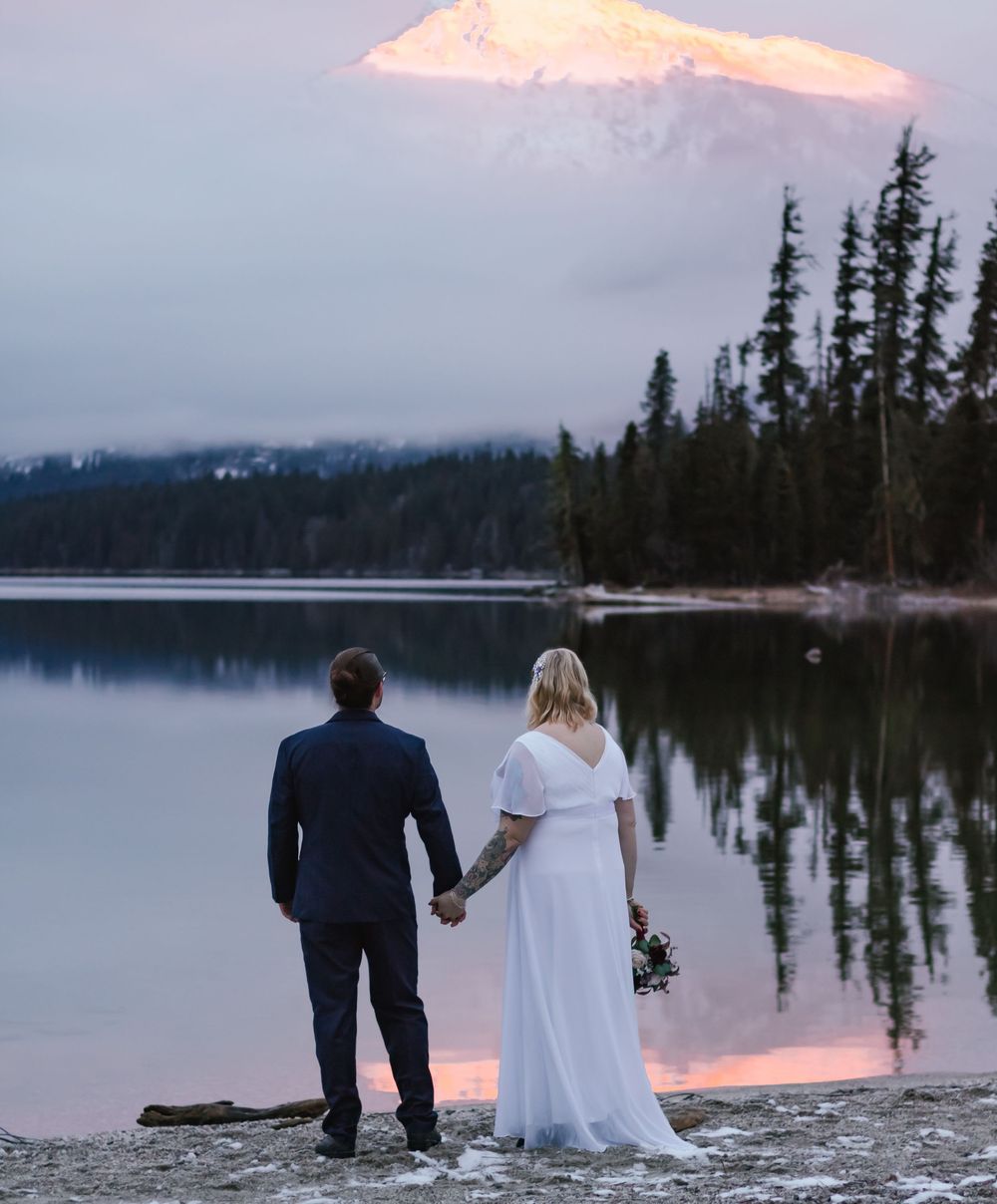 A couple stands at the edge of a tranquil lake surrounded by evergreens with a snow-capped mountain glowing in sunset light.