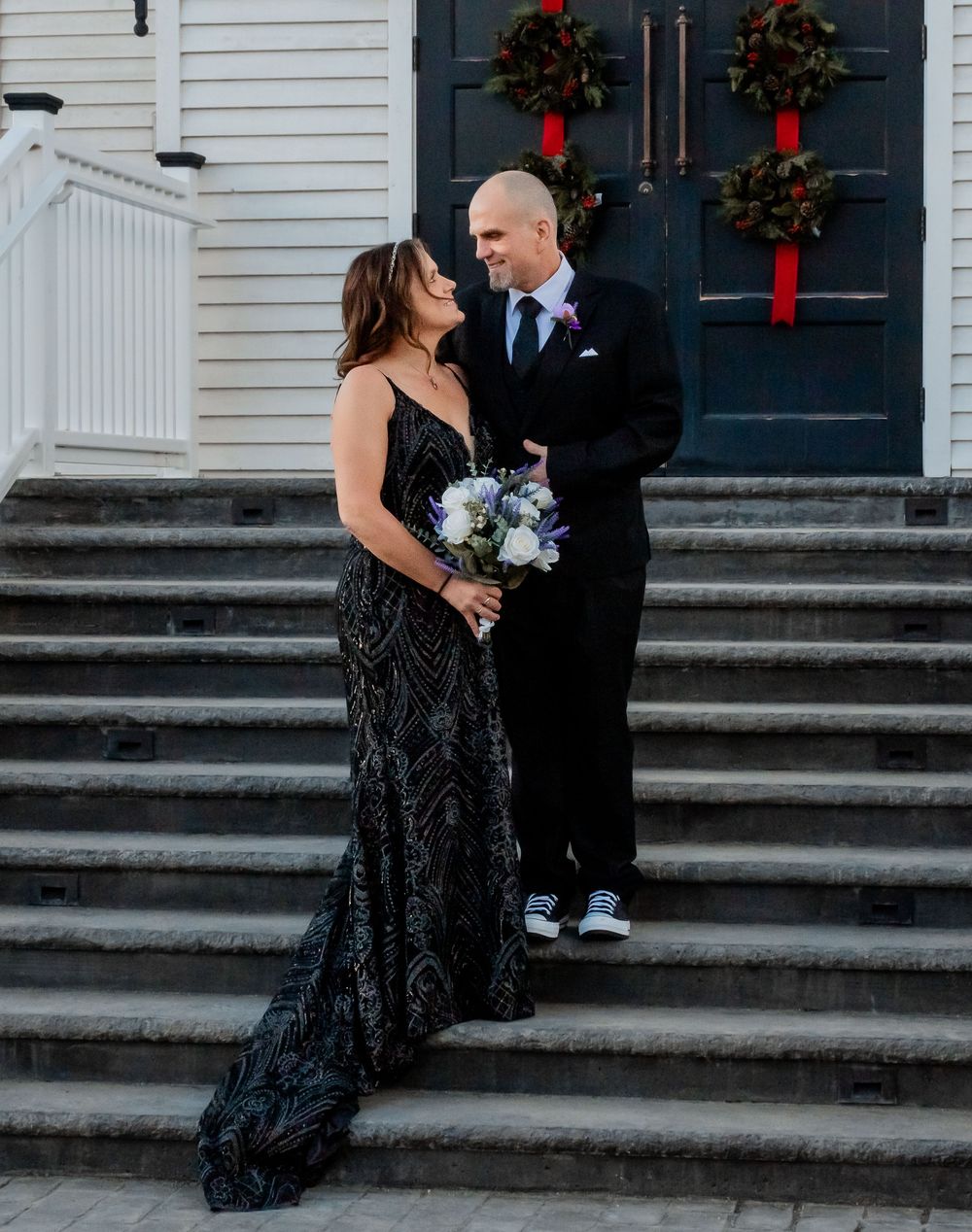 A couple in formal attire stands on church steps decorated with holiday wreaths during a winter wedding celebration.