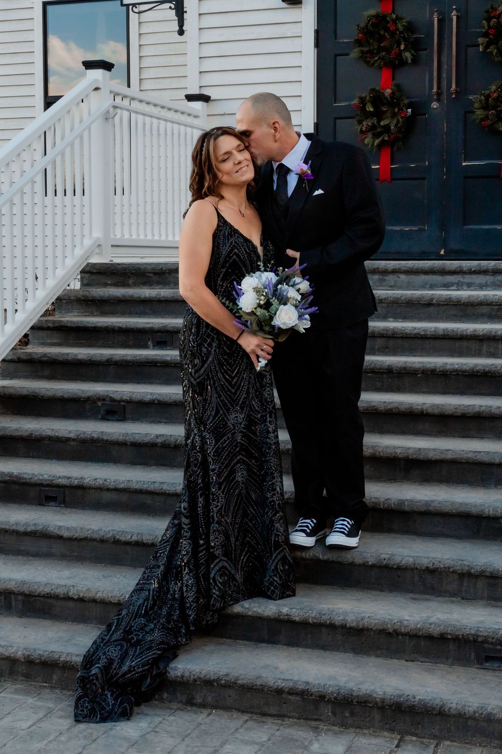Couple in formal attire embrace on white church steps with blue and white winter floral bouquet.