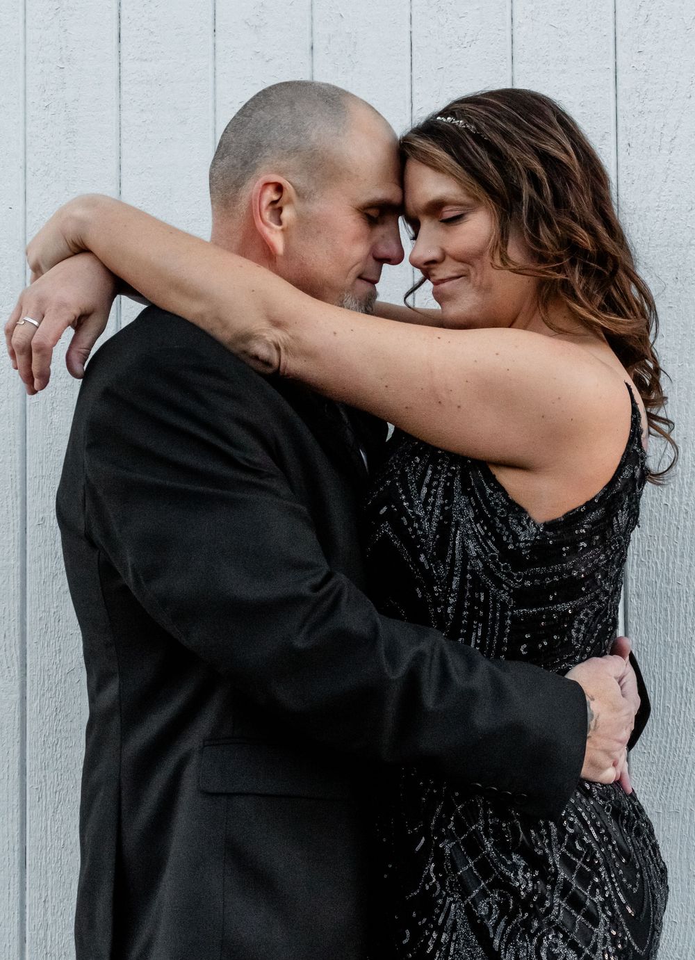Romantic couple embraces in formal attire against white wall, with her in sparkling black dress and him in dark suit.