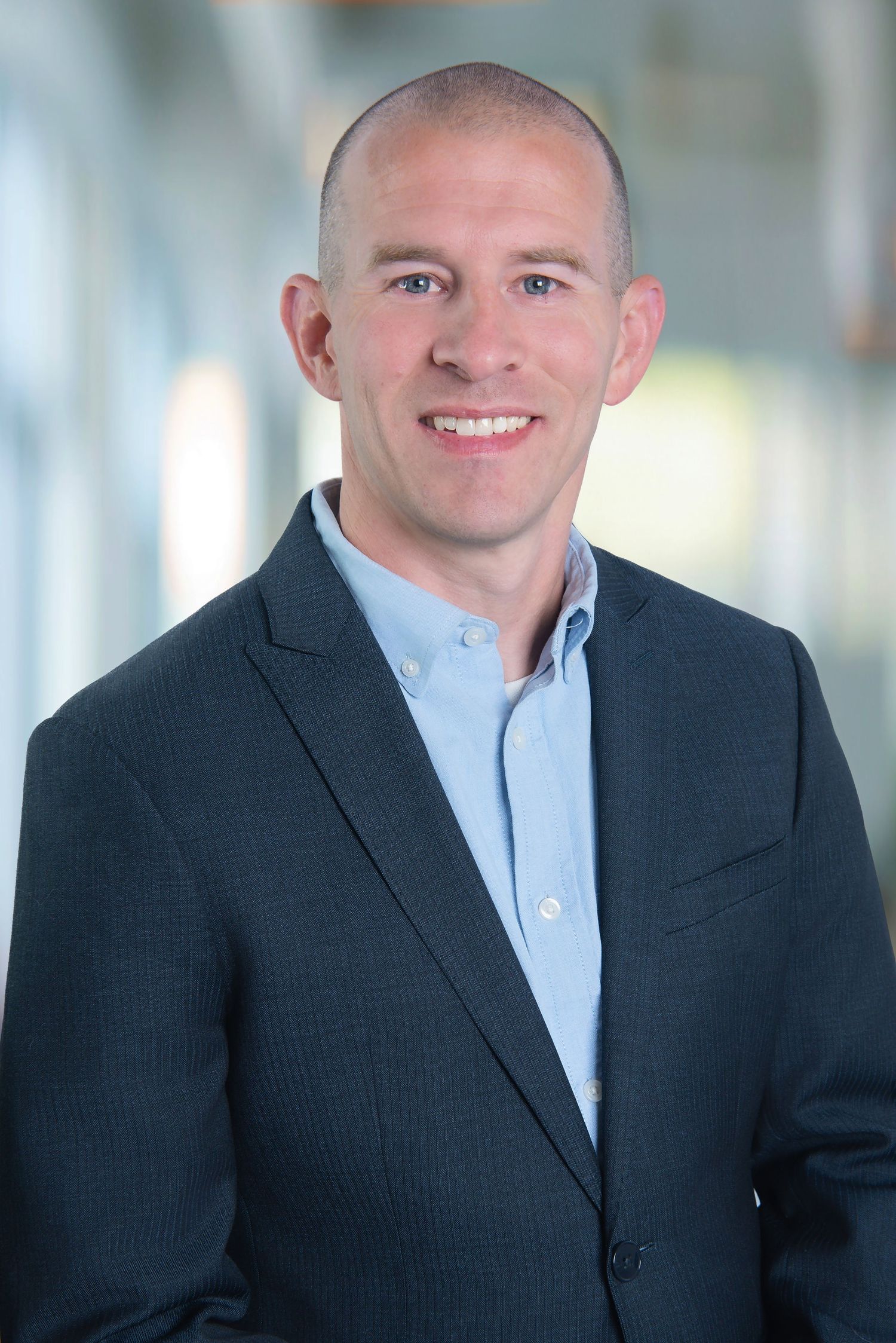 Professional headshot of a smiling man wearing a navy blazer and light blue button-up shirt against a blurred background.