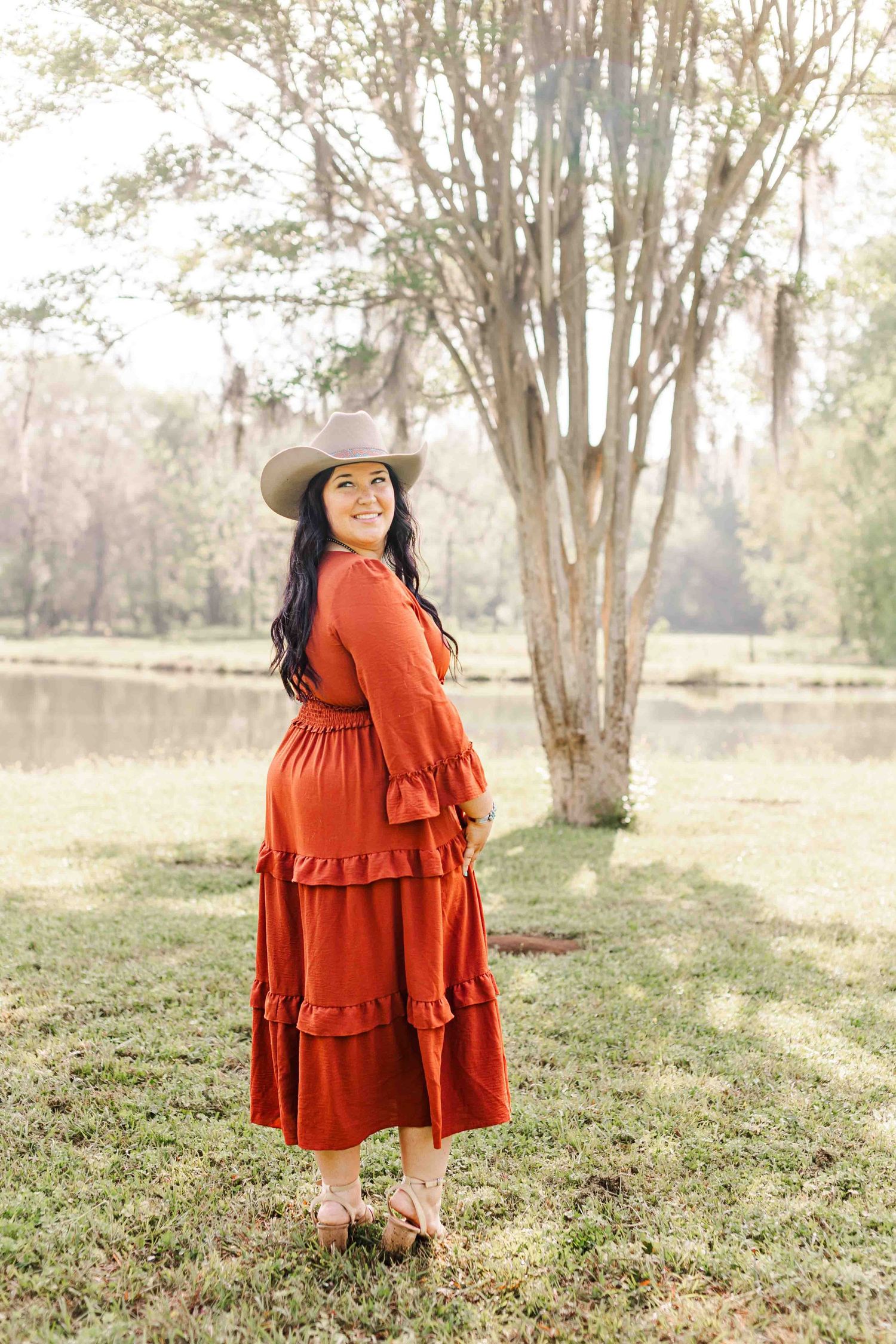 A woman in a rustic red tiered maxi dress and brown hat poses outdoors near a lake and trees.