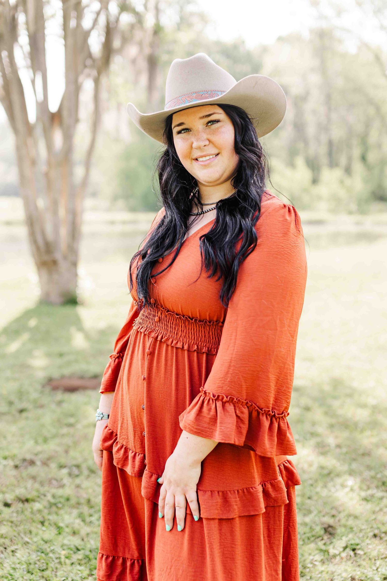 Person in rust-colored ruffled maxi dress and beige cowboy hat poses outdoors in a natural setting.