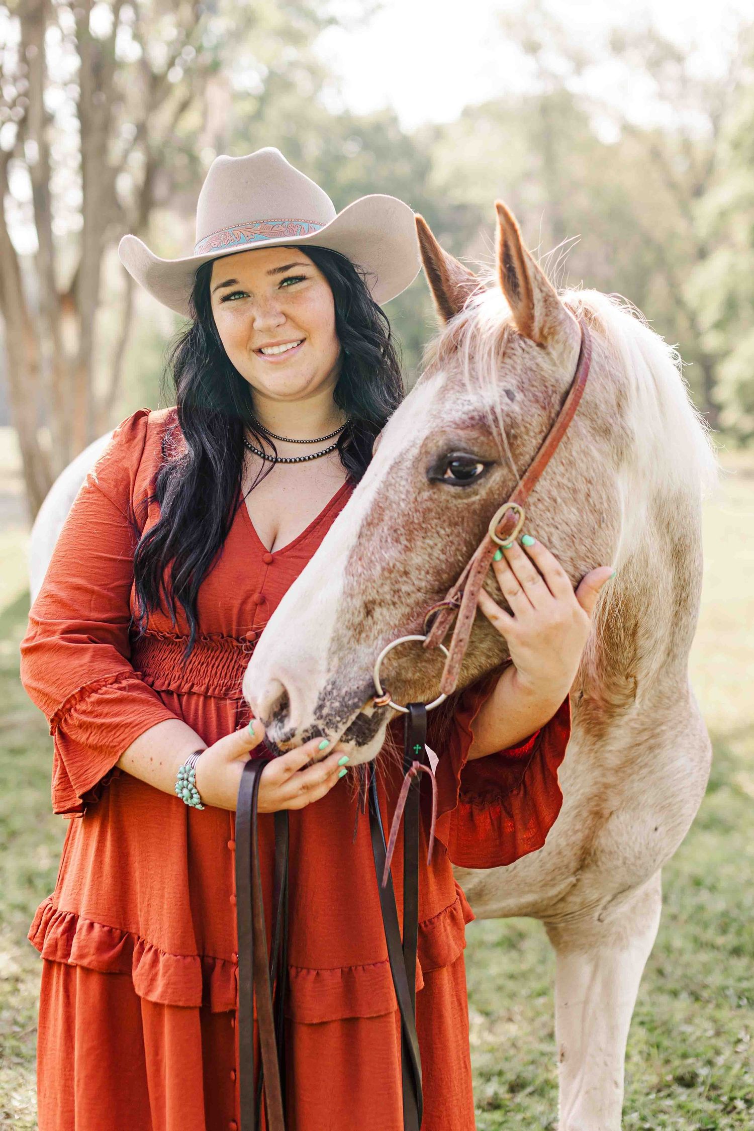 Someone in a red dress and beige cowboy hat stands with a beautiful white horse in a sunny outdoor setting.