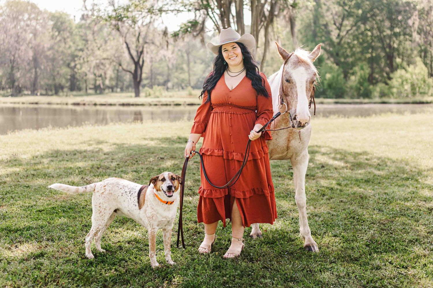 Person in rust dress poses with a white horse and spotted dog in a sunny field.