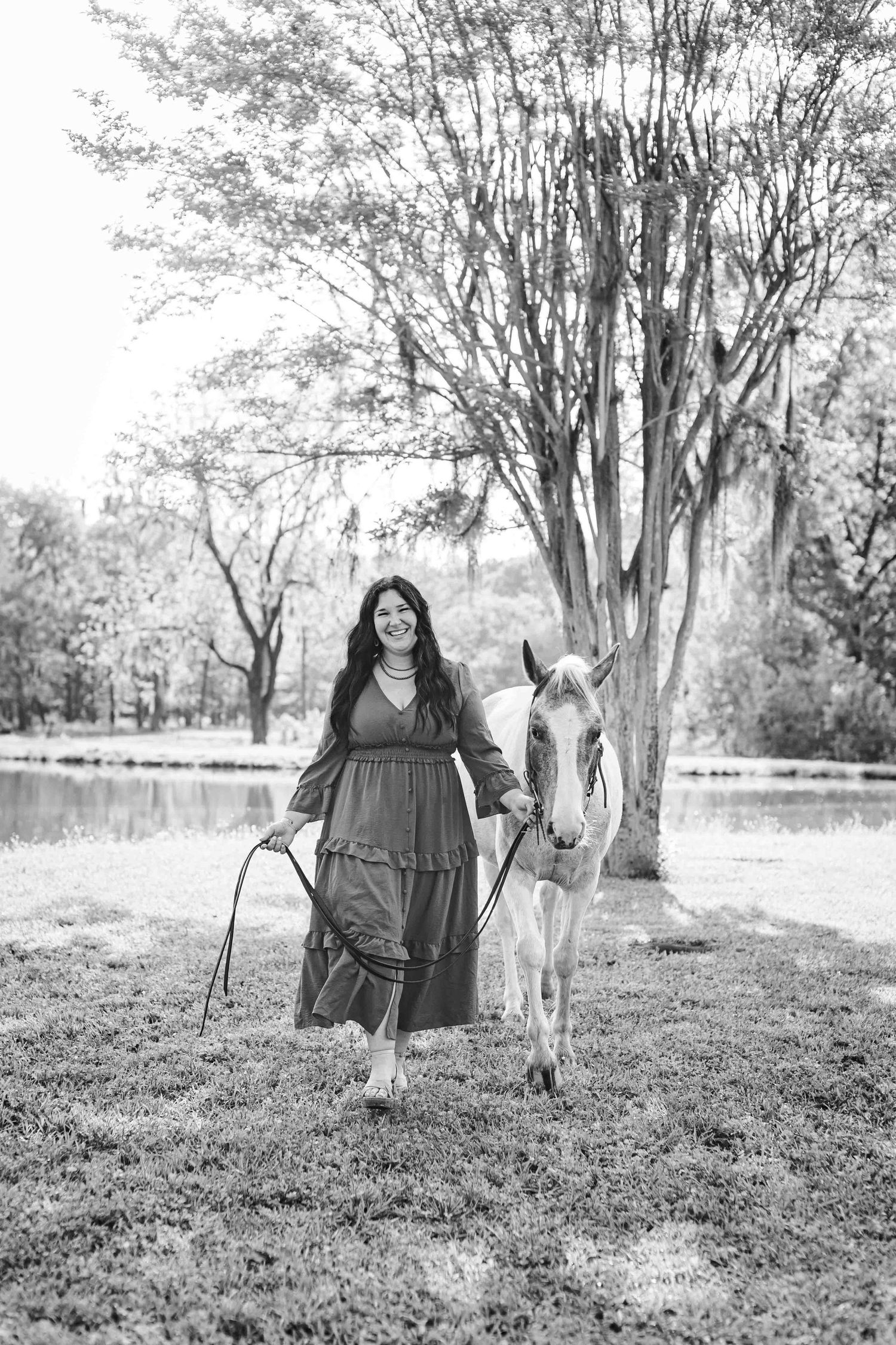 Black and white photo showing a person in a flowing dress walking with a white horse along a tranquil pond under tall trees.