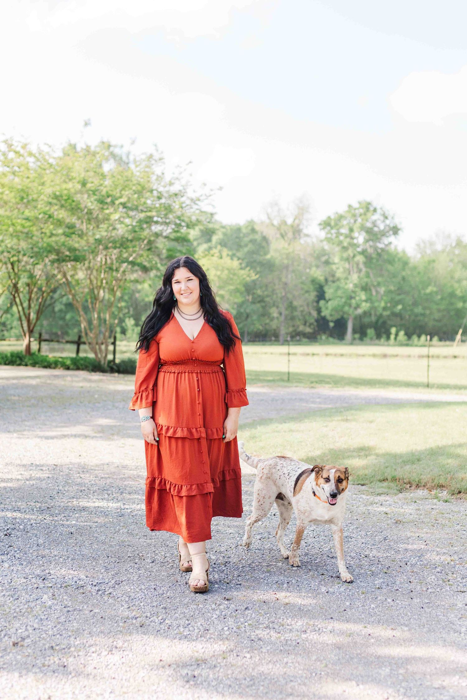 A person in a long rust-colored tiered dress walks along a gravel path with their spotted dog on a sunny day.