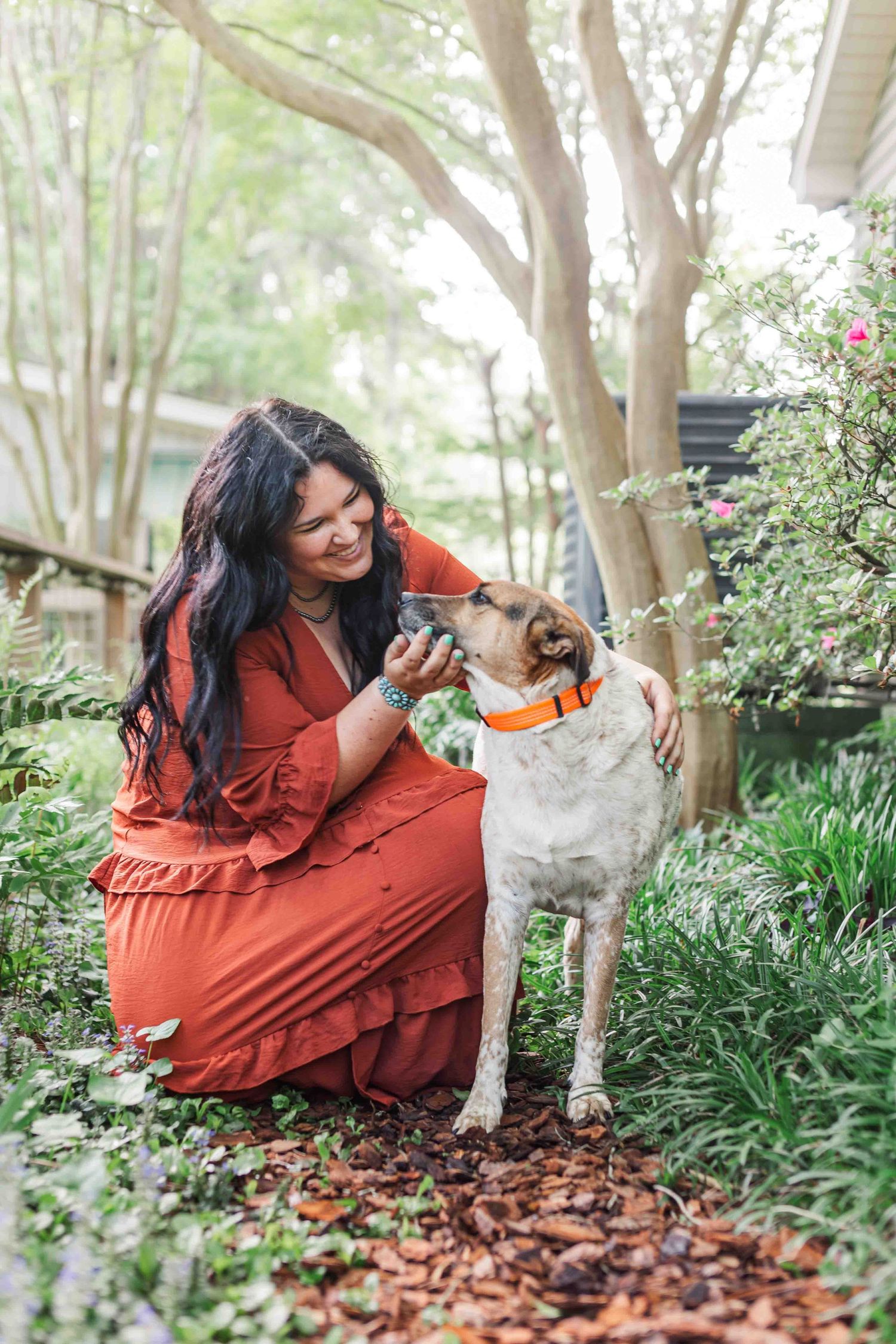 A person in a rust-colored dress shares a tender moment with their affectionate dog in a garden setting.