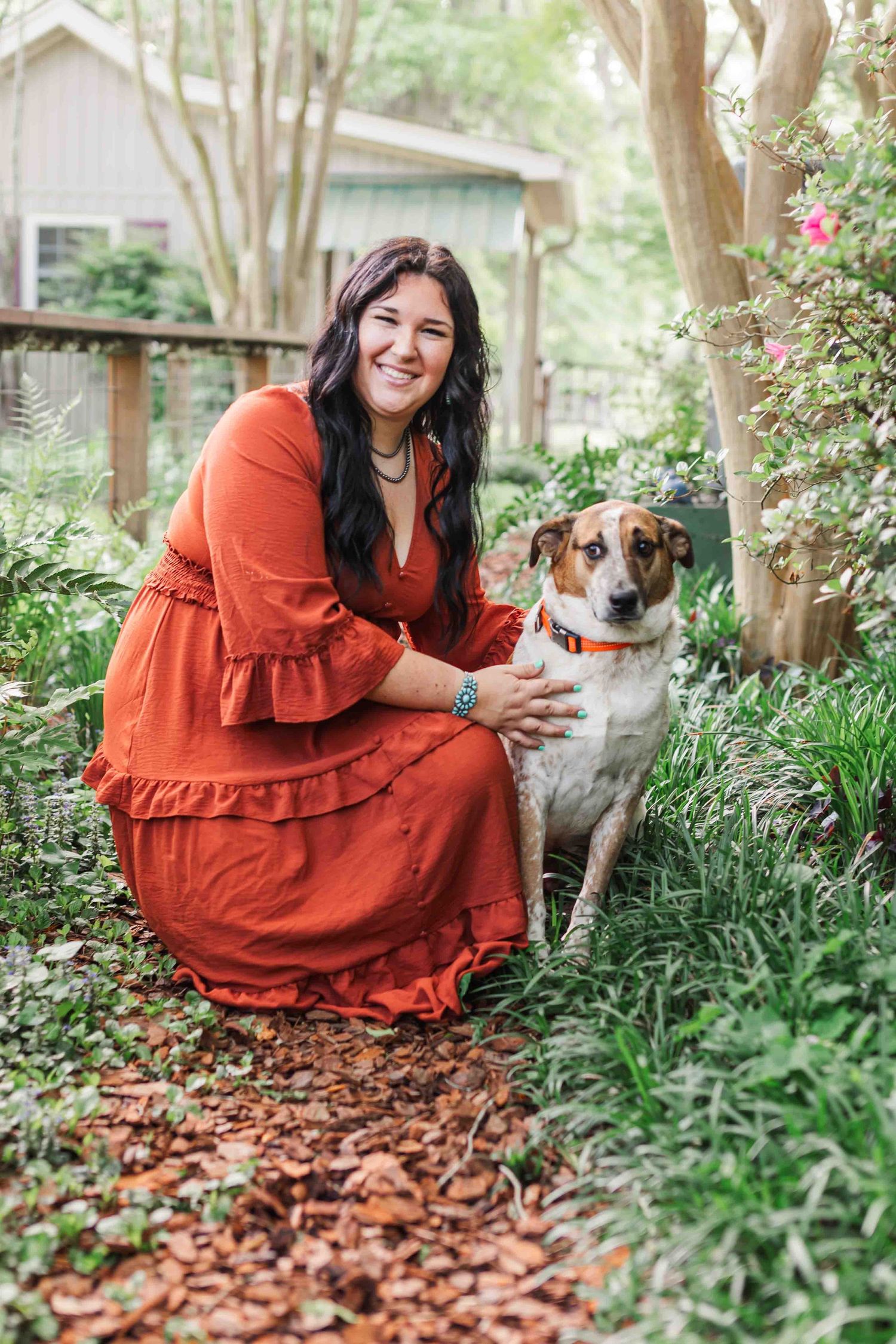 Someone in a flowy rust-colored dress kneels next to their mixed breed dog in a leafy garden setting.
