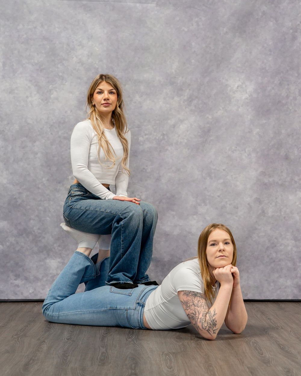 Two models in casual denim and white tops pose against gray backdrop for fashion shoot.