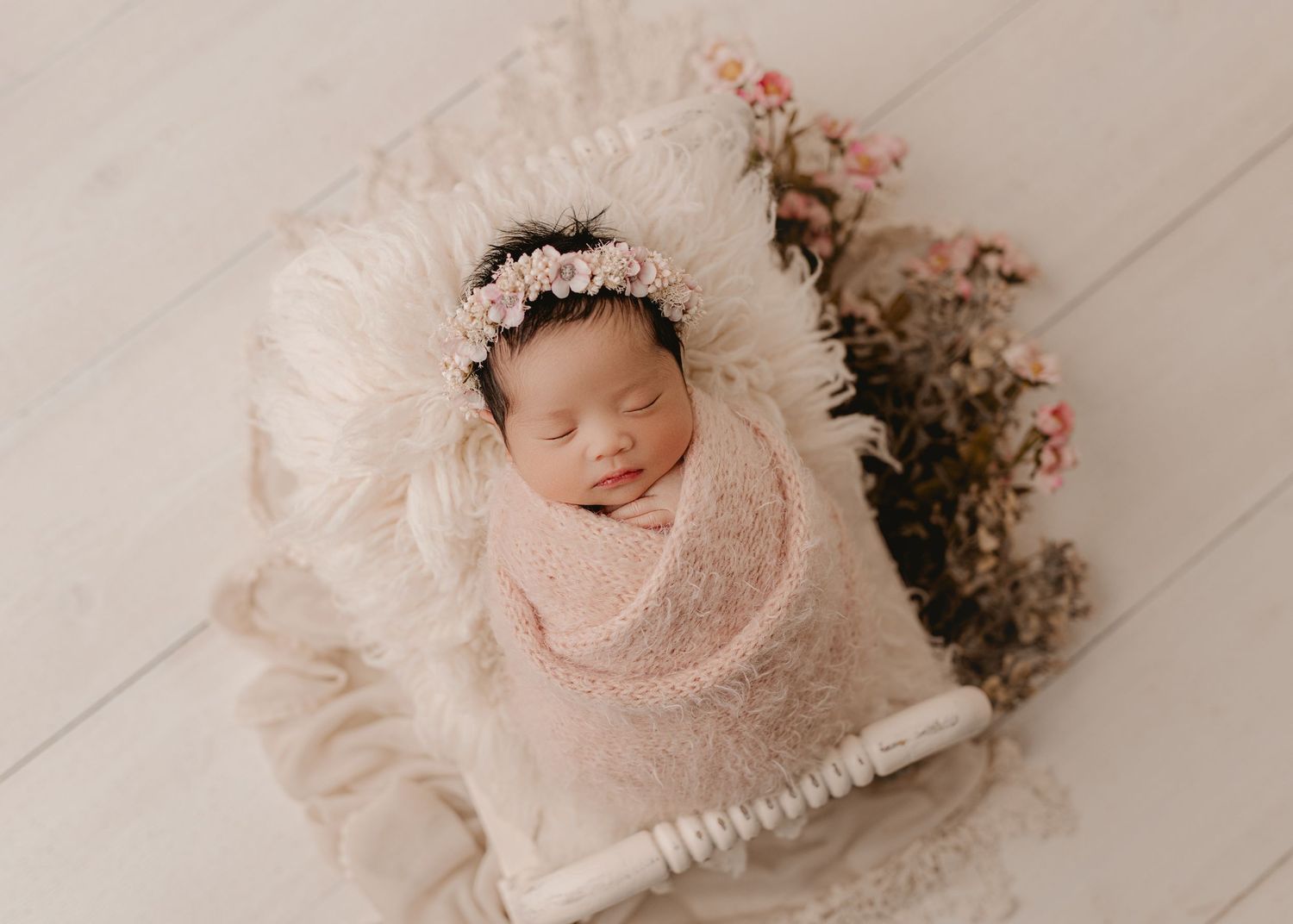 Sleeping newborn wrapped in soft pink swaddle with floral decor and white bonnet on white backdrop. Studio newborn photography MI