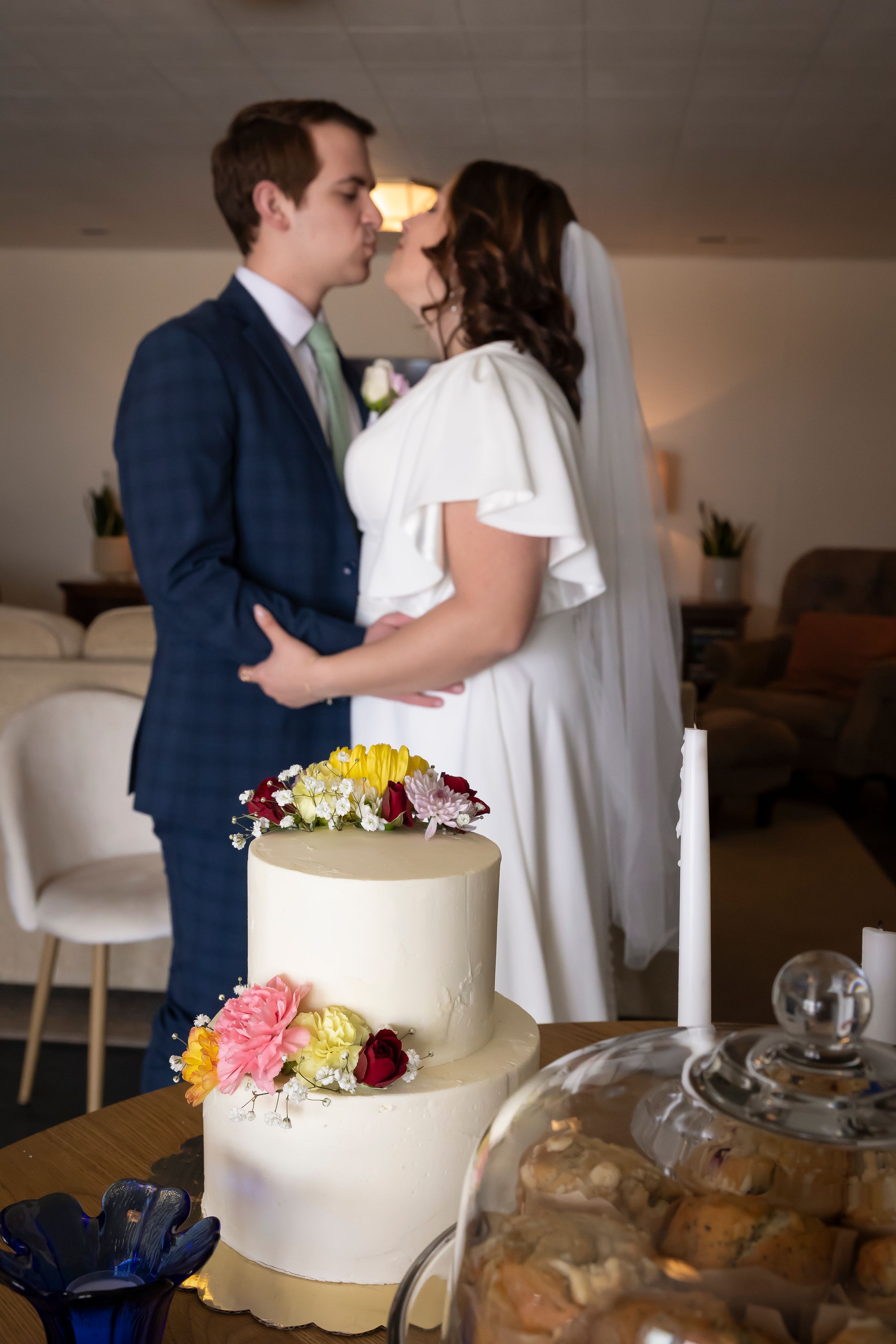 2 tier wedding cake with off white fondant and fresh spring flowers made by Crystal with Rose Hill Bakery with bride and groom in background