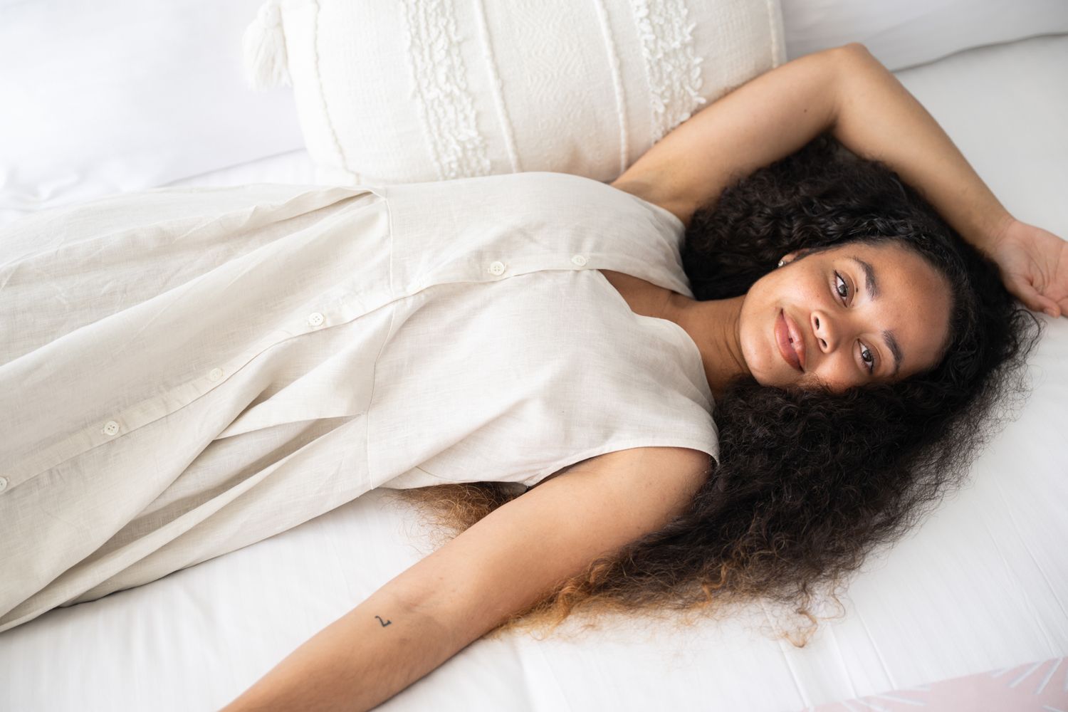 Woman with curly dark hair relaxes on white bedding in soft natural lighting.