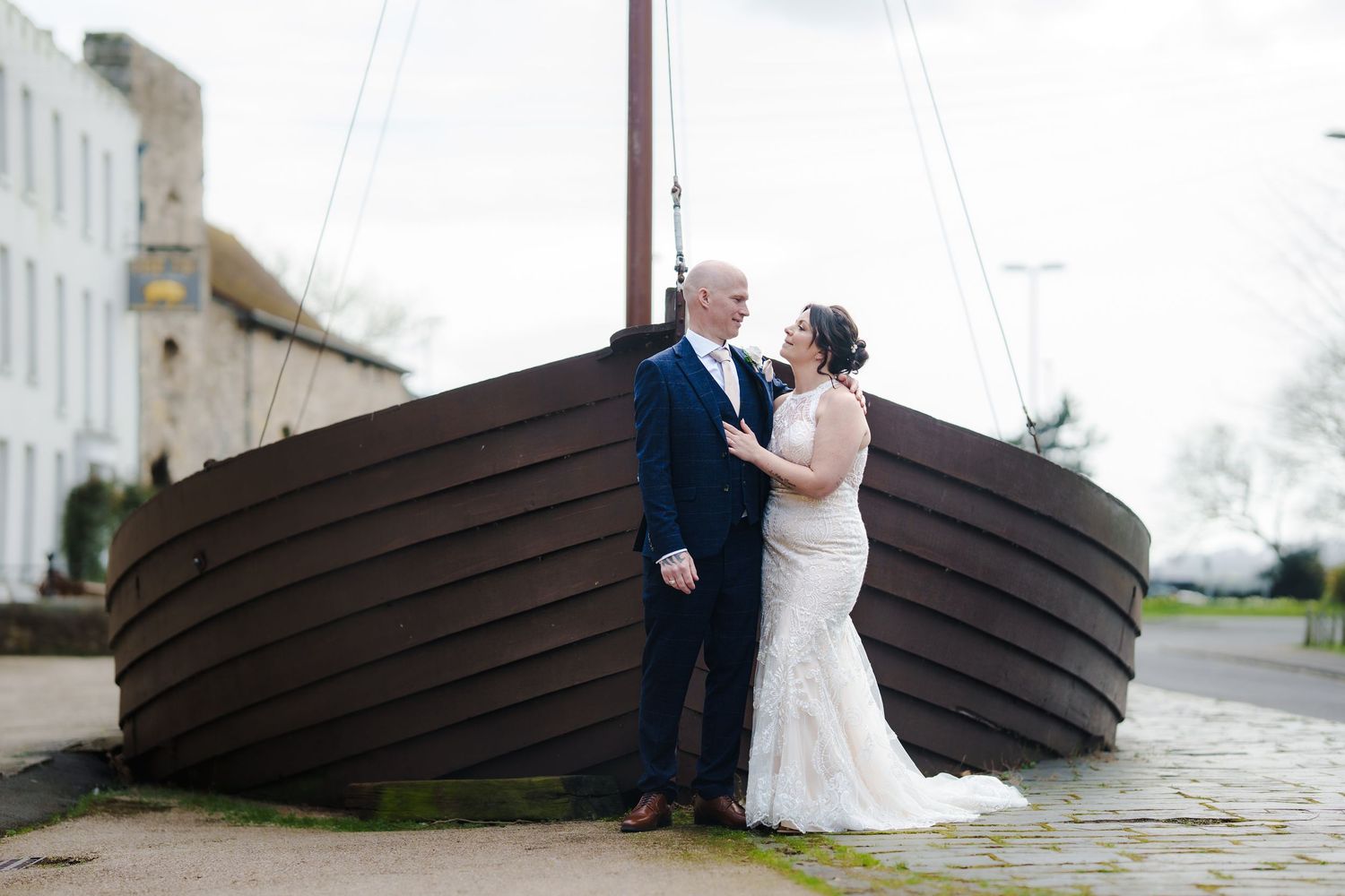 A couple in wedding attire embraces by a large wooden boat sculpture along a waterfront pathway.