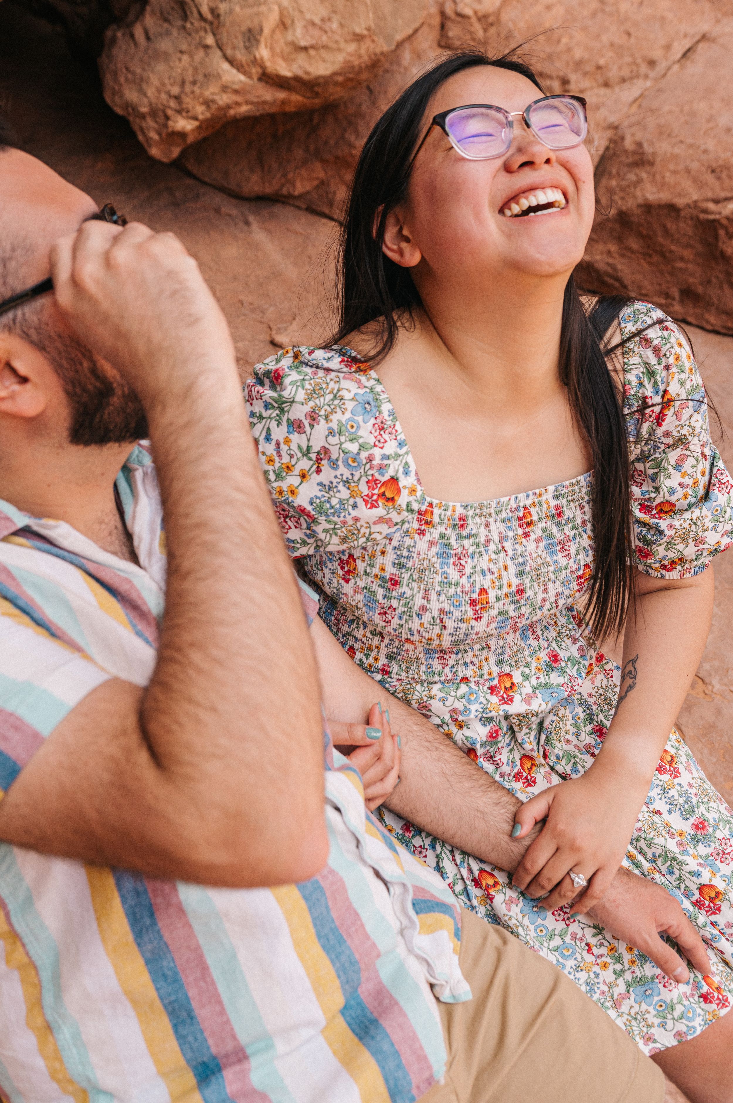 A female and male sitting on rocks at Arches National Park. The woman is wearing a white dress with a floral print and is laughing.