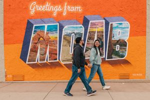 A female and male walk across a mural in Moab. They are looking at each other and laughing. The mural says Greetings from Moab.