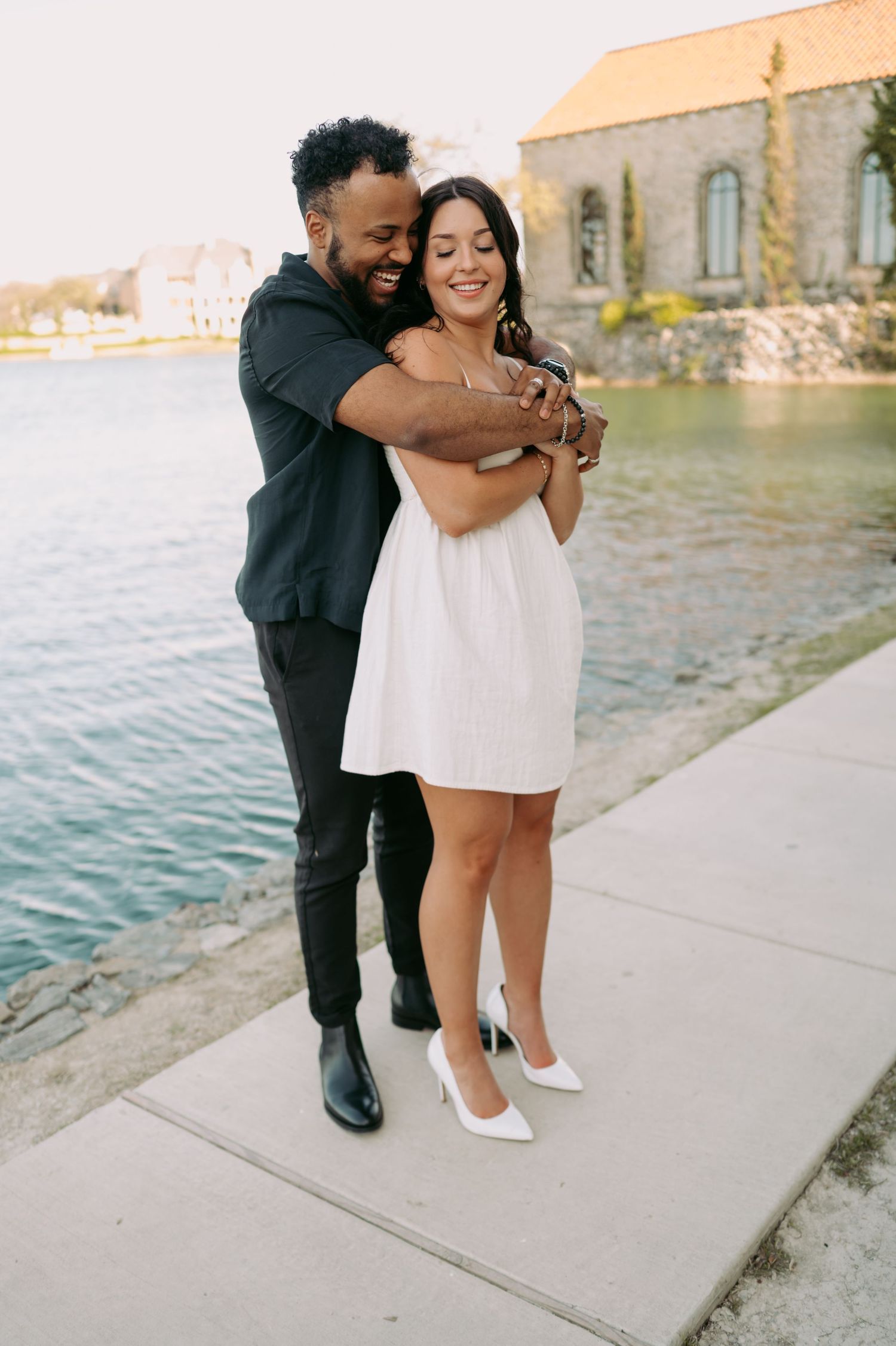 A couple embraces on a waterfront path with a stone church in the background.