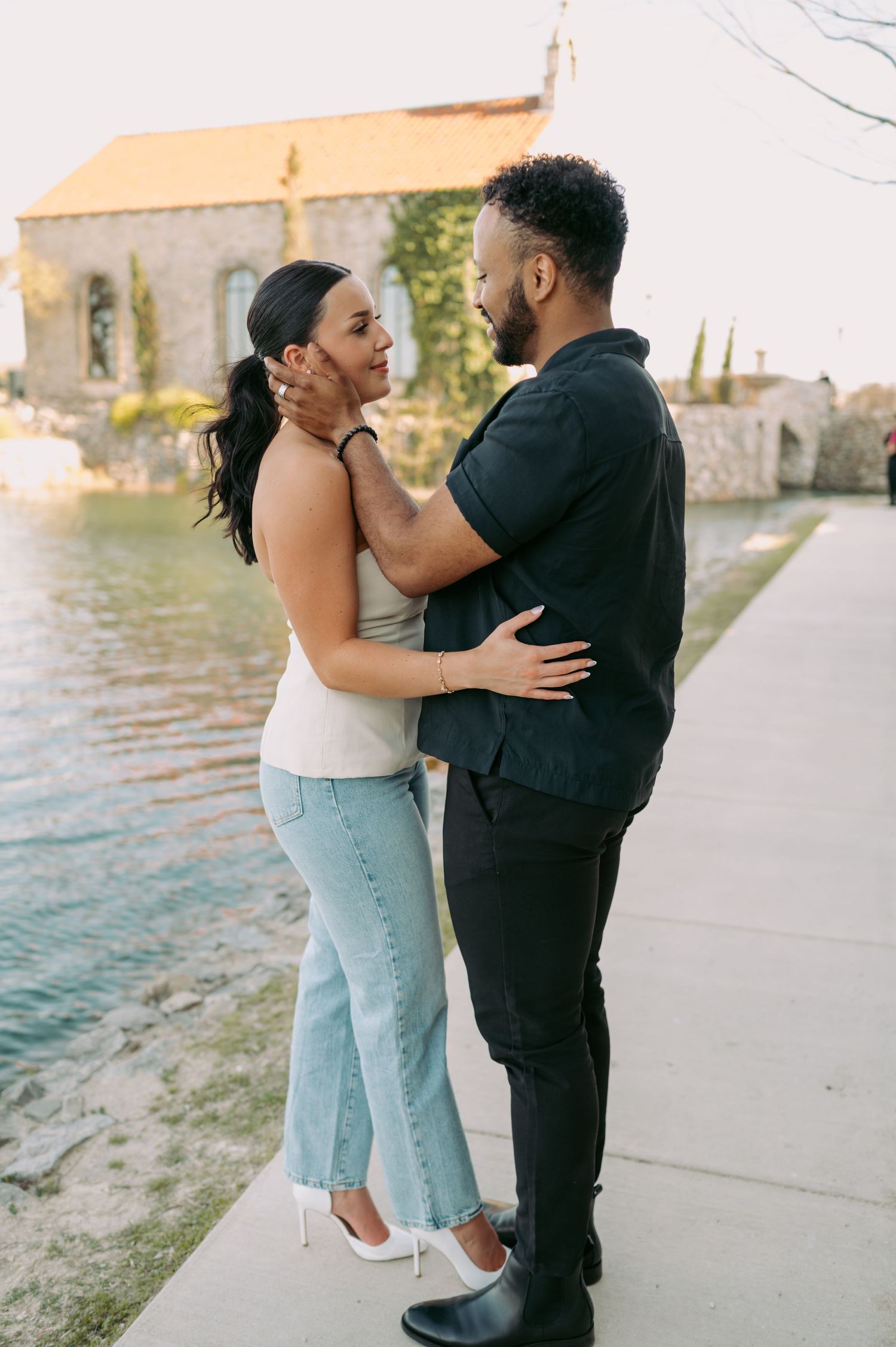 A romantic couple embraces near a stone church and waterfront during a sunset photoshoot.