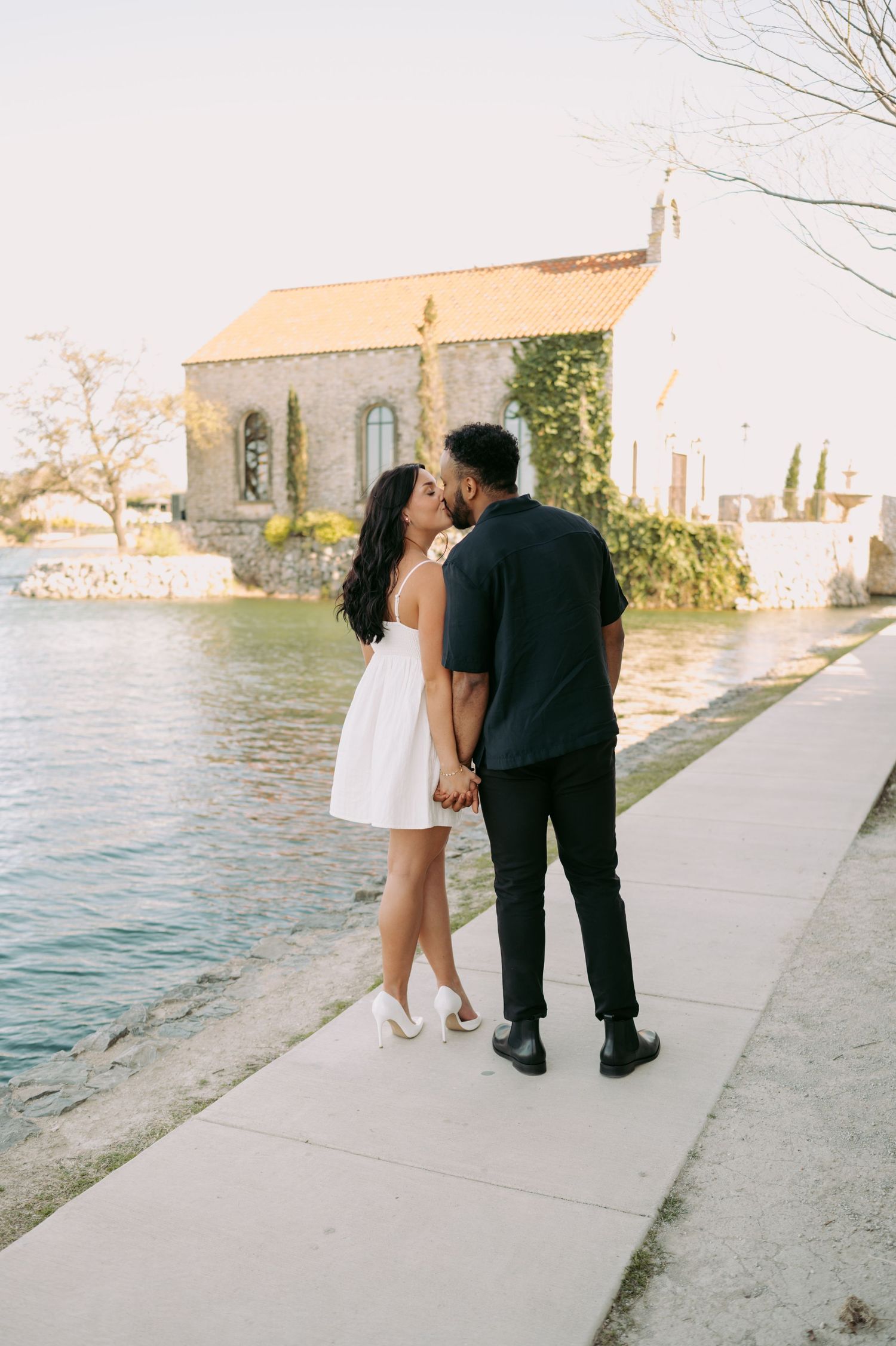 Couple sharing intimate moment by water with stone chapel in background.