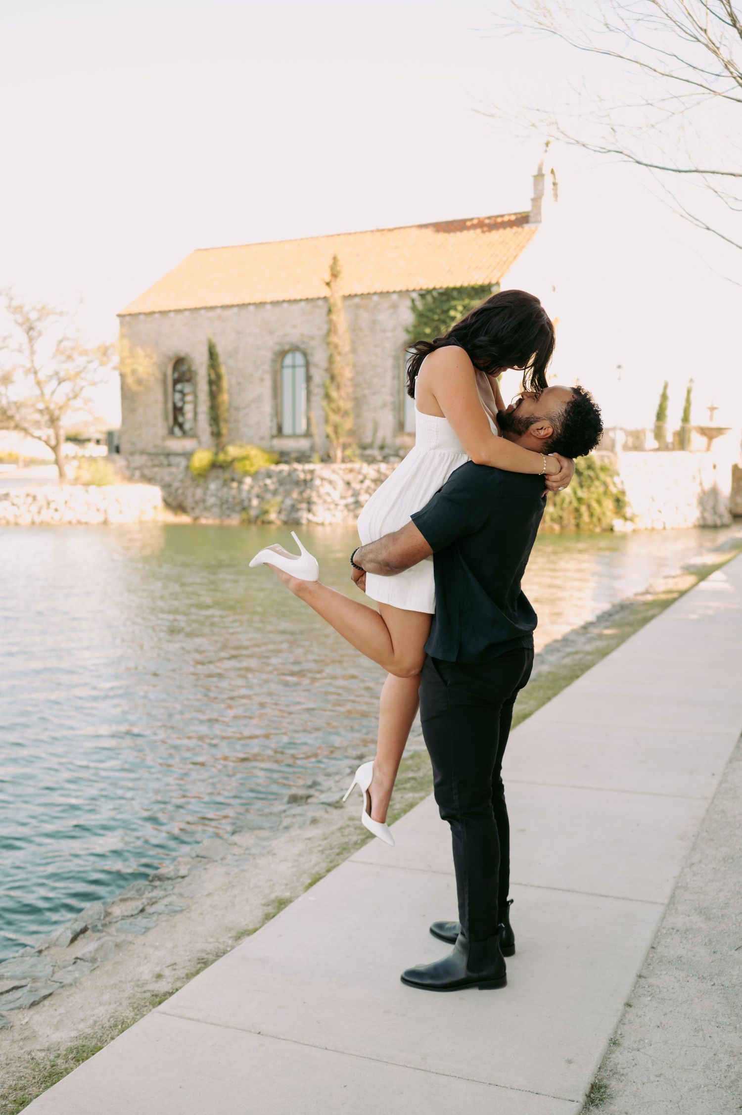 Romantic couple embracing by waterfront with chapel in background.