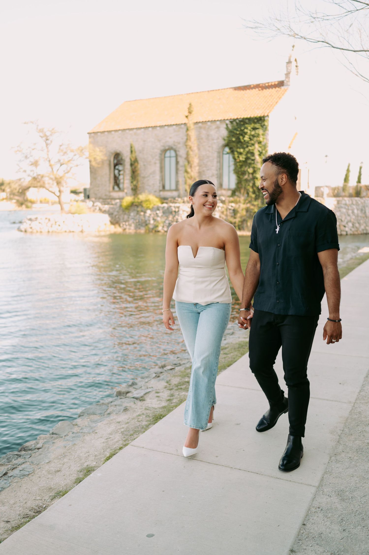 Happy couple walking hand in hand along waterfront path near stone chapel.