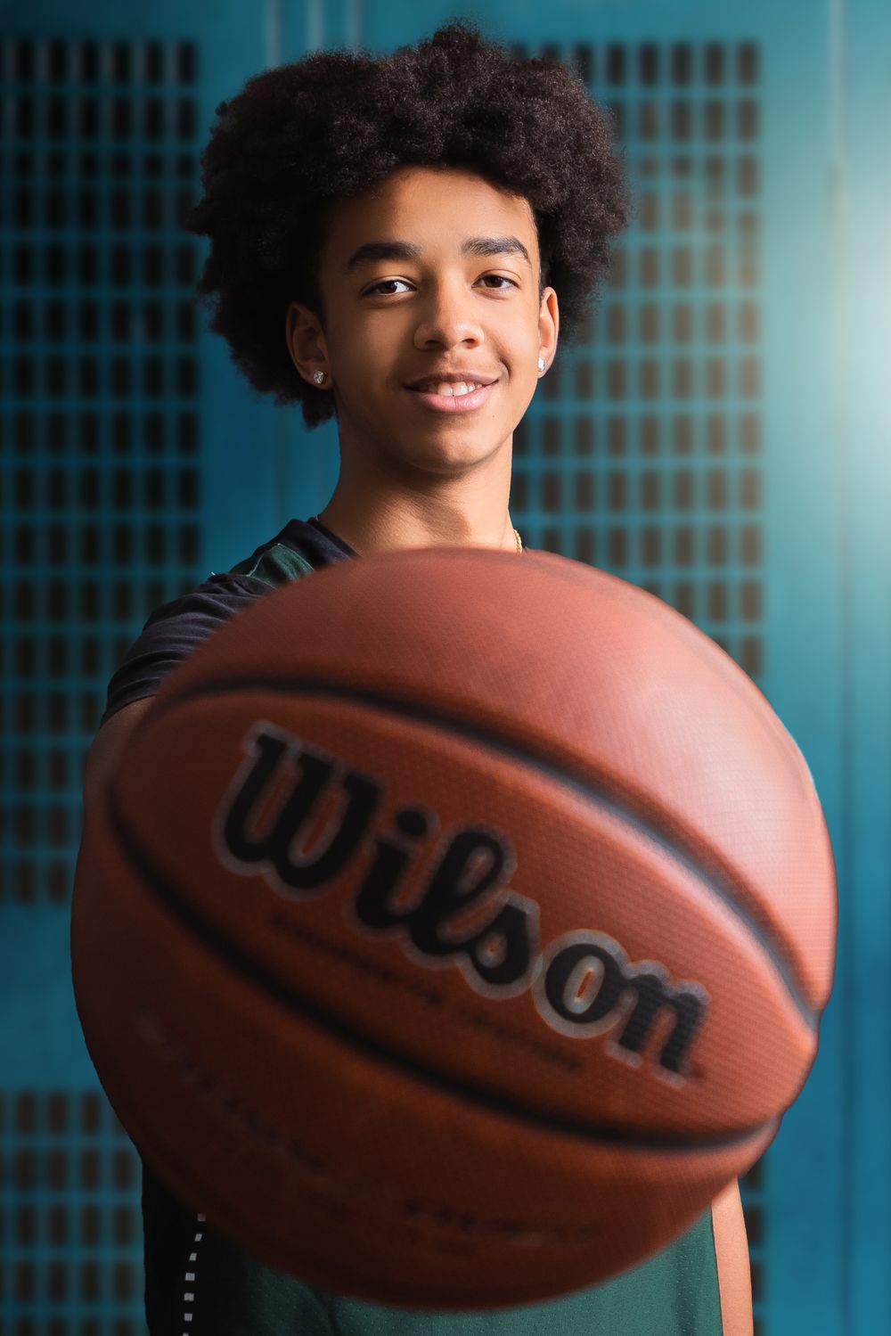 A young athlete with curly hair holds a Wilson basketball against a turquoise background.