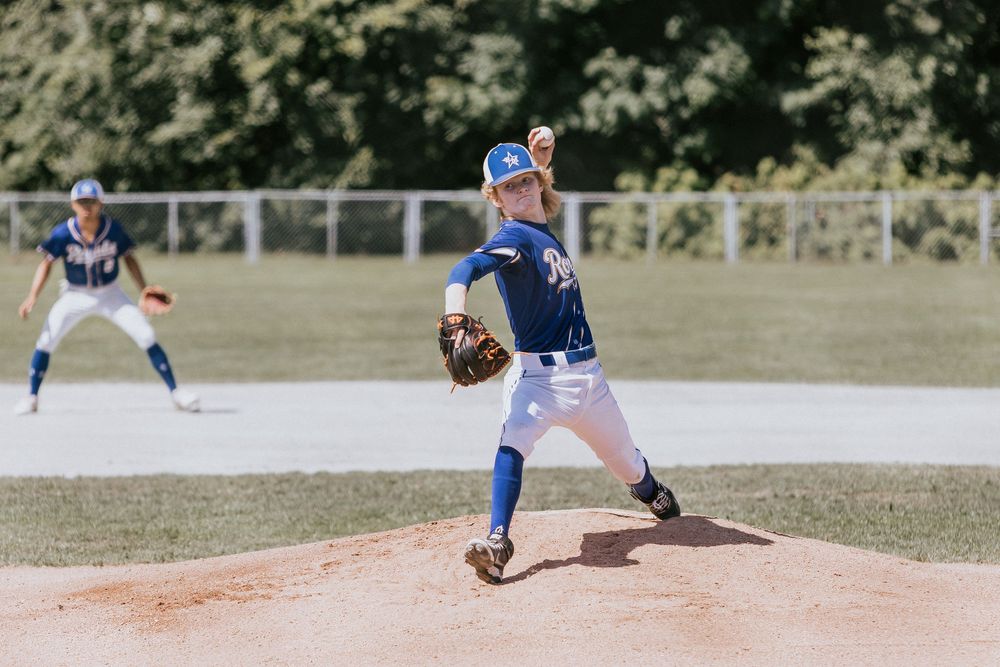 Youth baseball pitcher in blue uniform mid-delivery on dirt mound during daytime game.