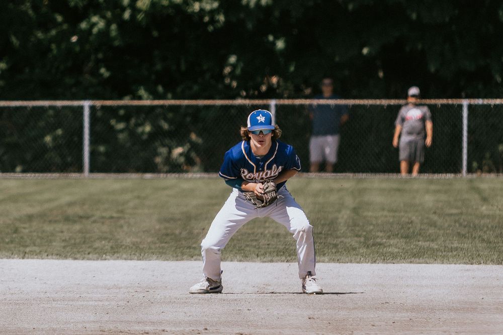 Baseball player in blue cap crouches in ready position on baseball base during outdoor game.