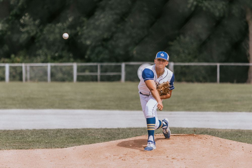 A youth baseball pitcher wearing a white and blue uniform stands in mid-pitching motion on a dirt mound.