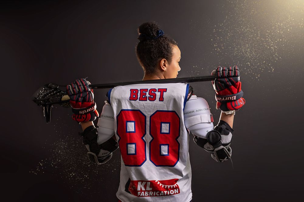 Athlete in white jersey with number 88 holds lacrosse stick across shoulders in dramatic lighting with floating particles.