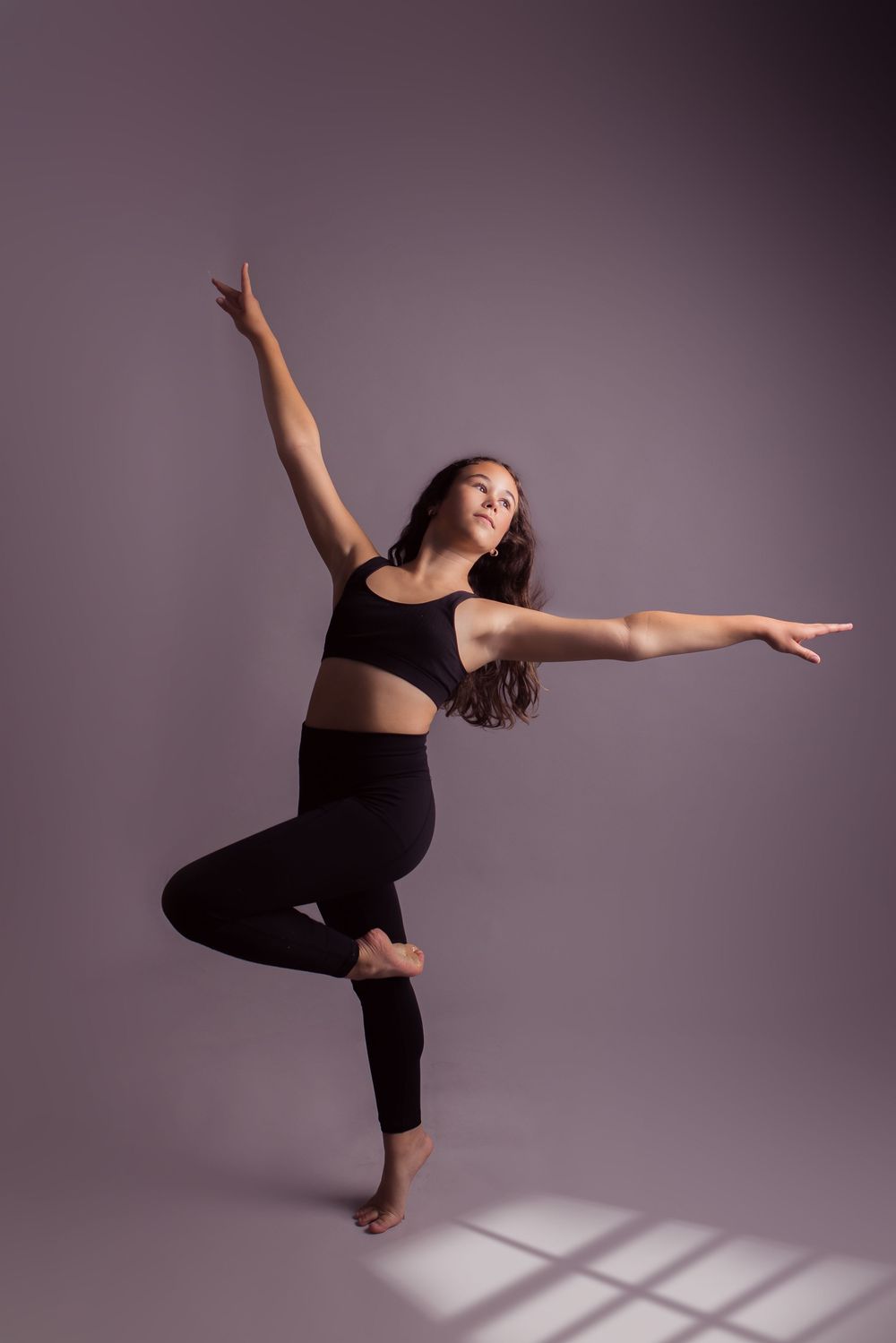A dancer in black athletic wear performs a graceful balancing pose in a studio with dramatic lighting and shadows.
