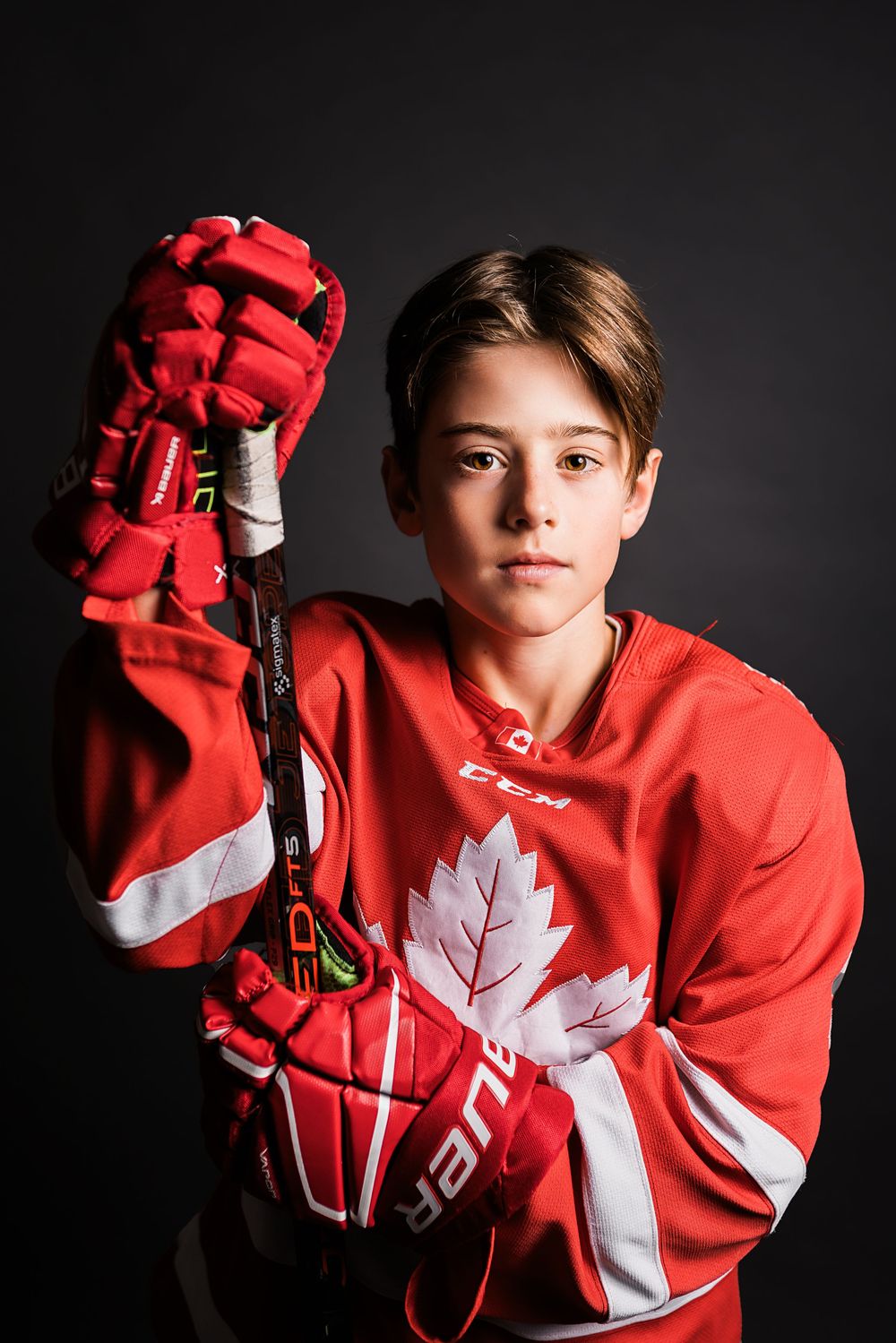 A hockey player poses in a red Team Canada jersey with maple leaf logo and matching red gloves against dark background.