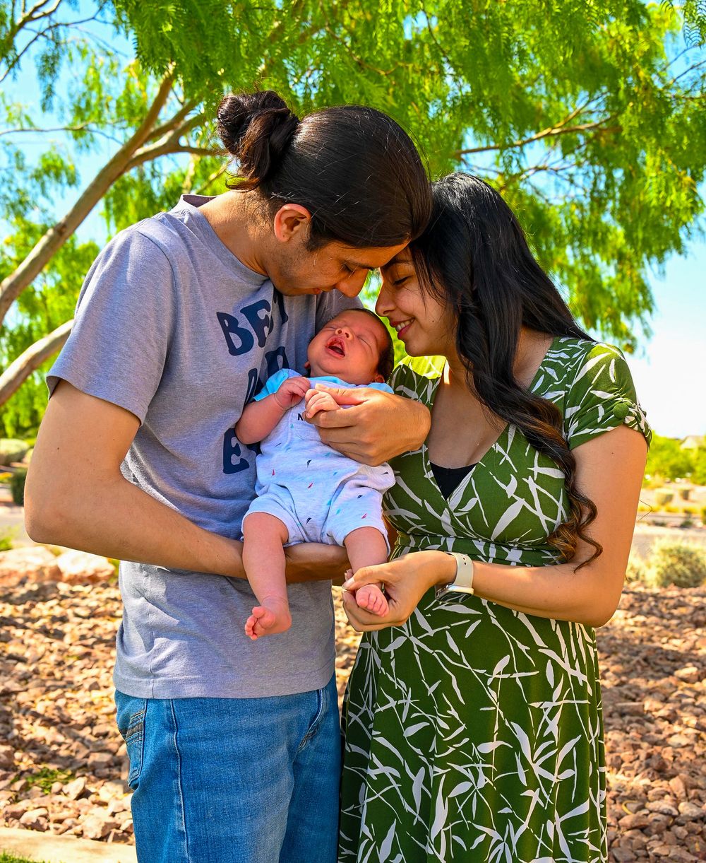 Young parents kissing their newborn baby outdoors surrounded by green trees.