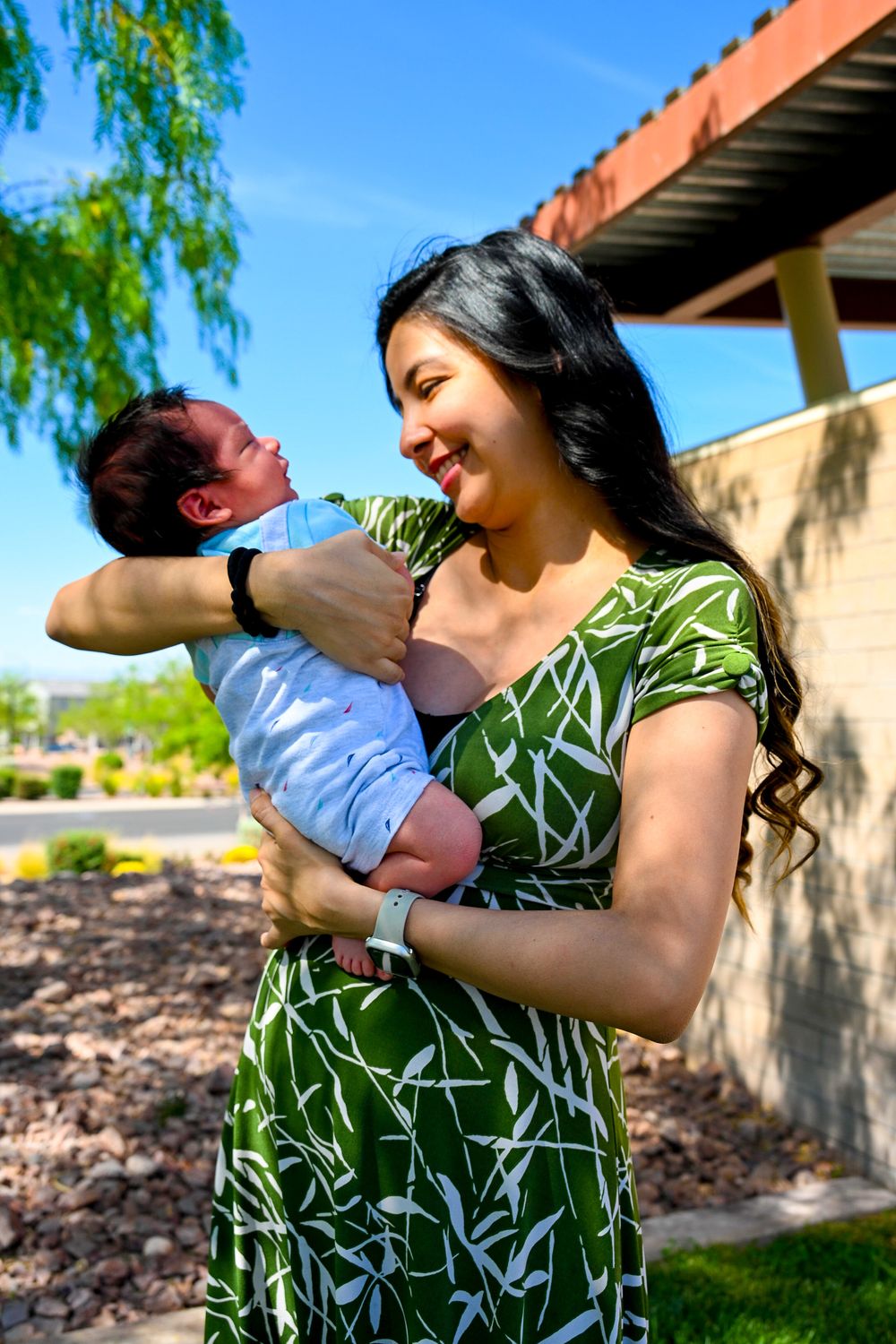 Smiling young mother in green dress lovingly holding her newborn baby outdoors.