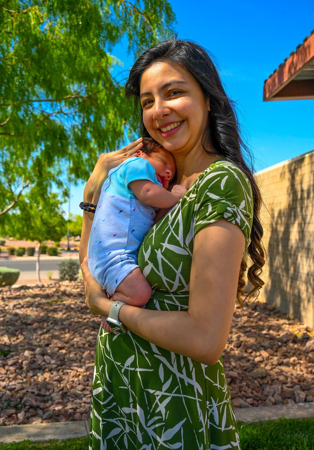 Joyful young mother in green floral dress holding her sleeping newborn baby outdoors.