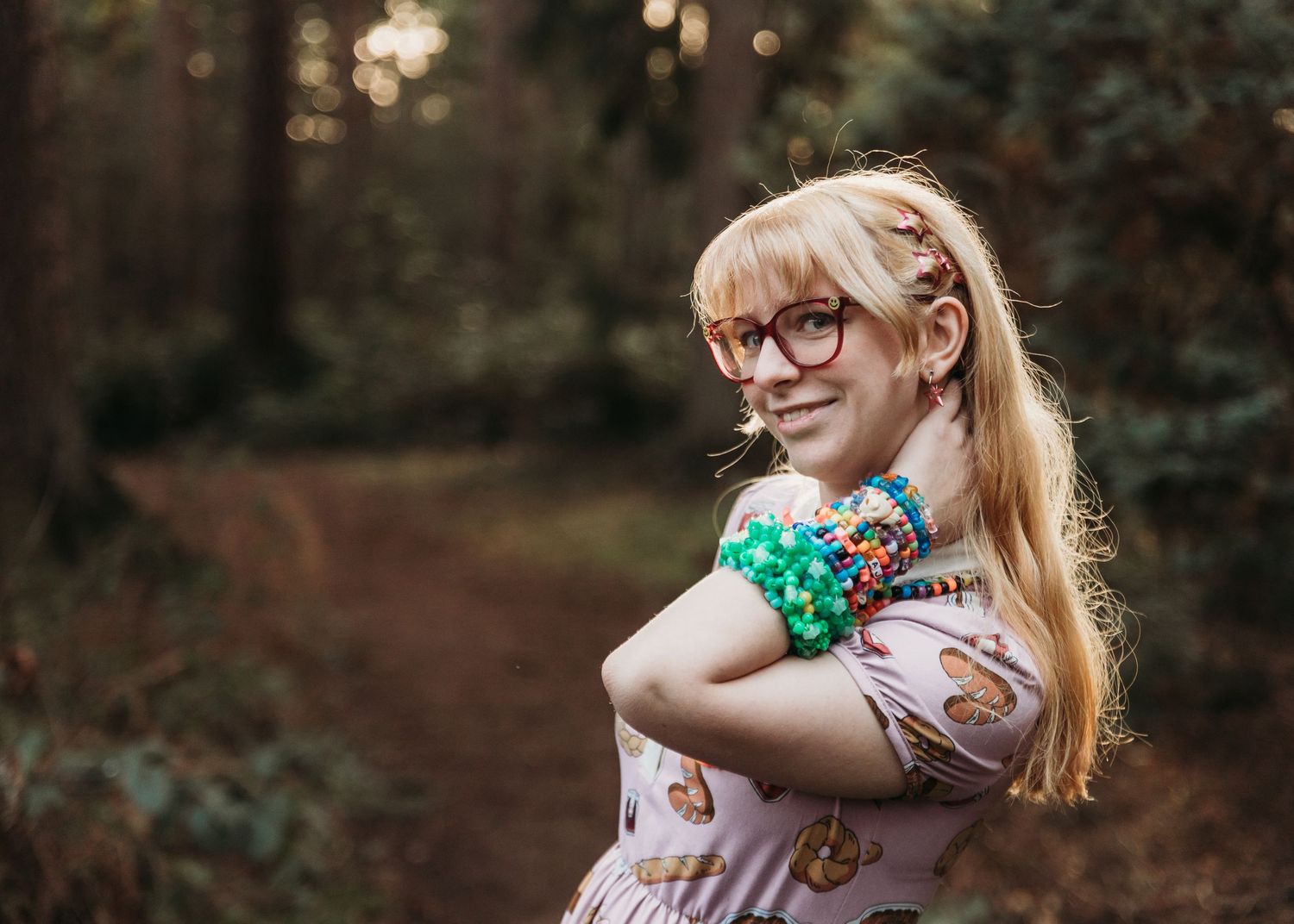 A person in glasses and a floral dress poses in a dreamy forest setting at golden hour.