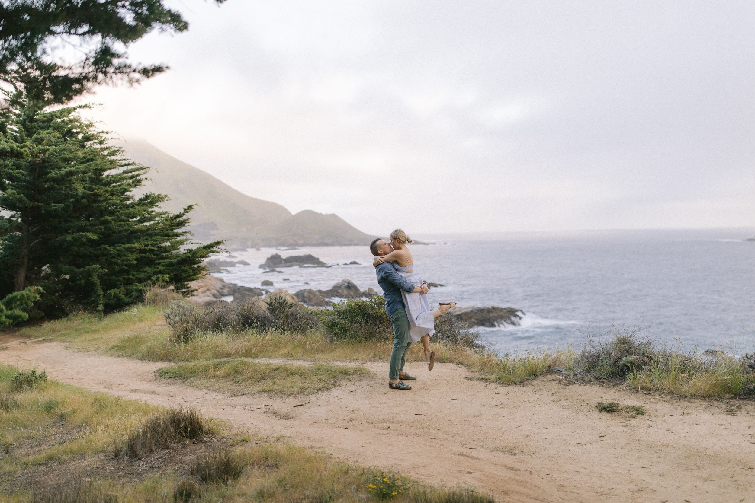 A person stands on a coastal cliff trail overlooking the Pacific Ocean on a cloudy day near rugged coastline and evergreens.