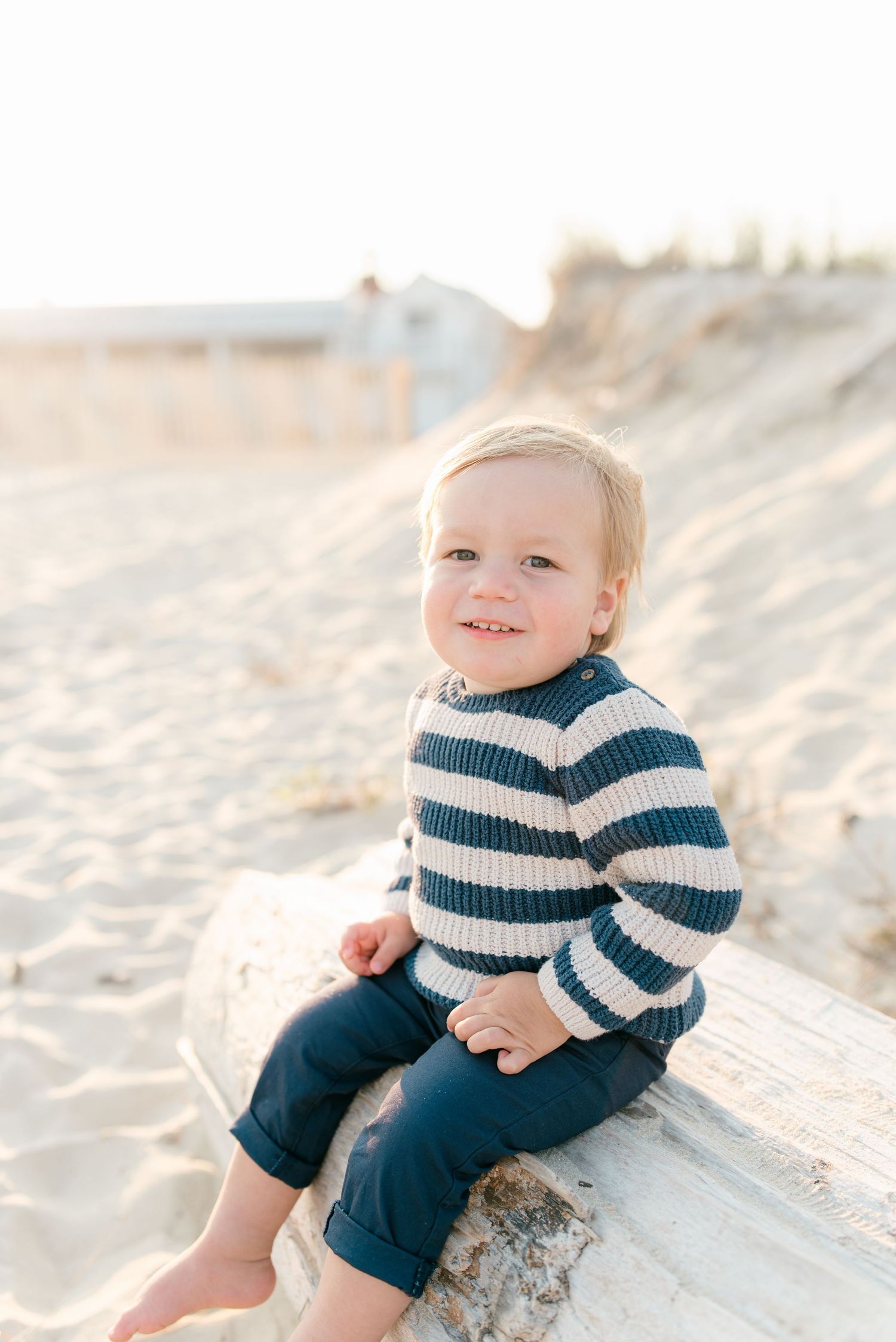 18 month old boy sits on a piece of driftwood on the beach smiling with the sun behind him.