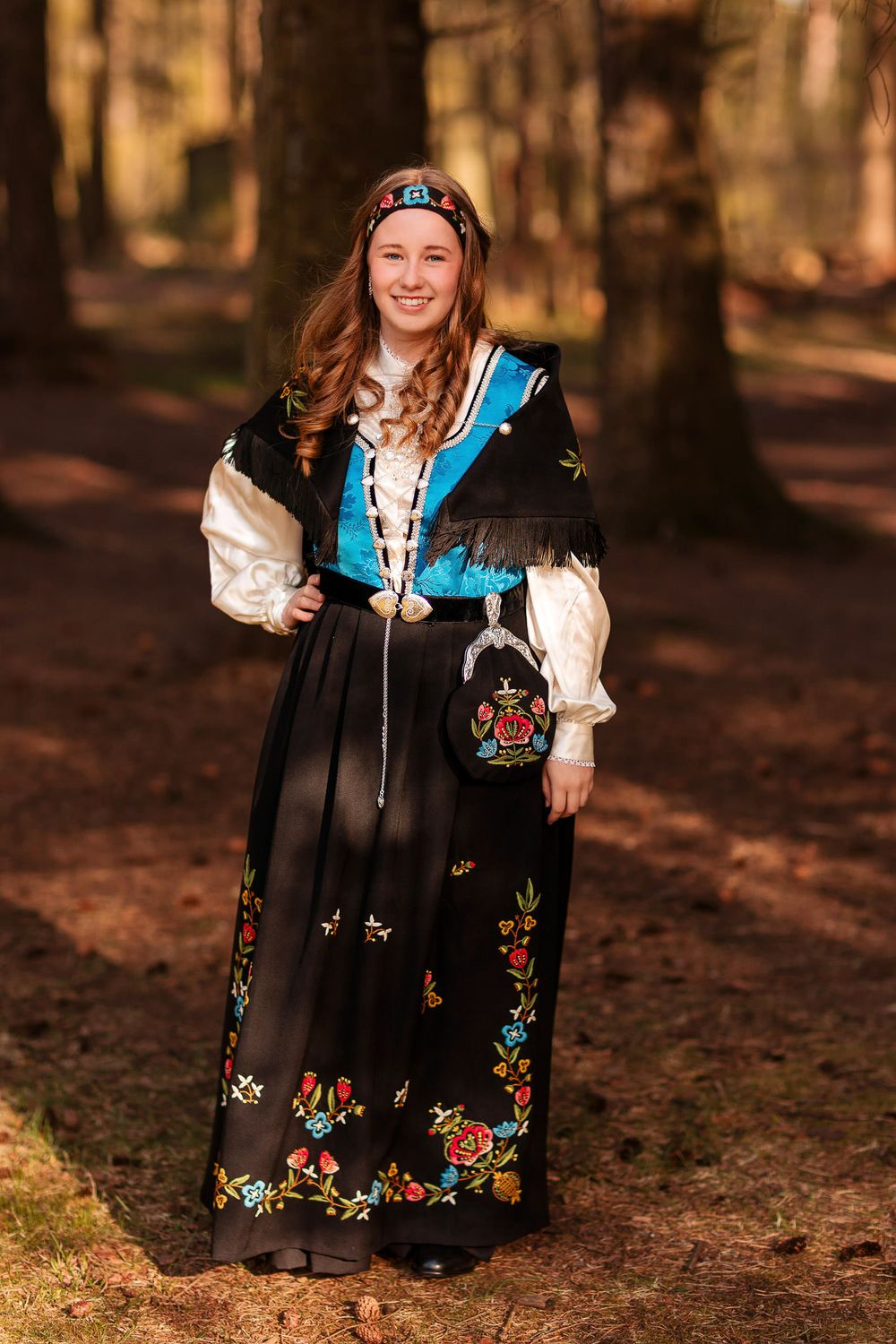 Nydelig konfirmant i bunad fra Jelsa med blå vest og hårbånd. Hun står i en skog på Ølberg i Sola kommune.