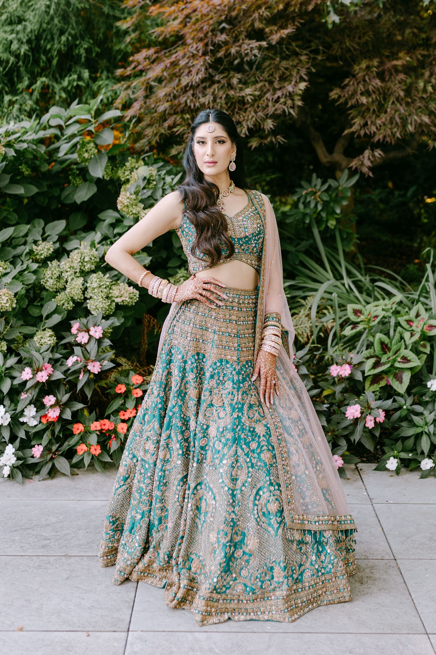 Traditional Indian bridal lehenga in turquoise and gold with intricate embroidery against garden backdrop.