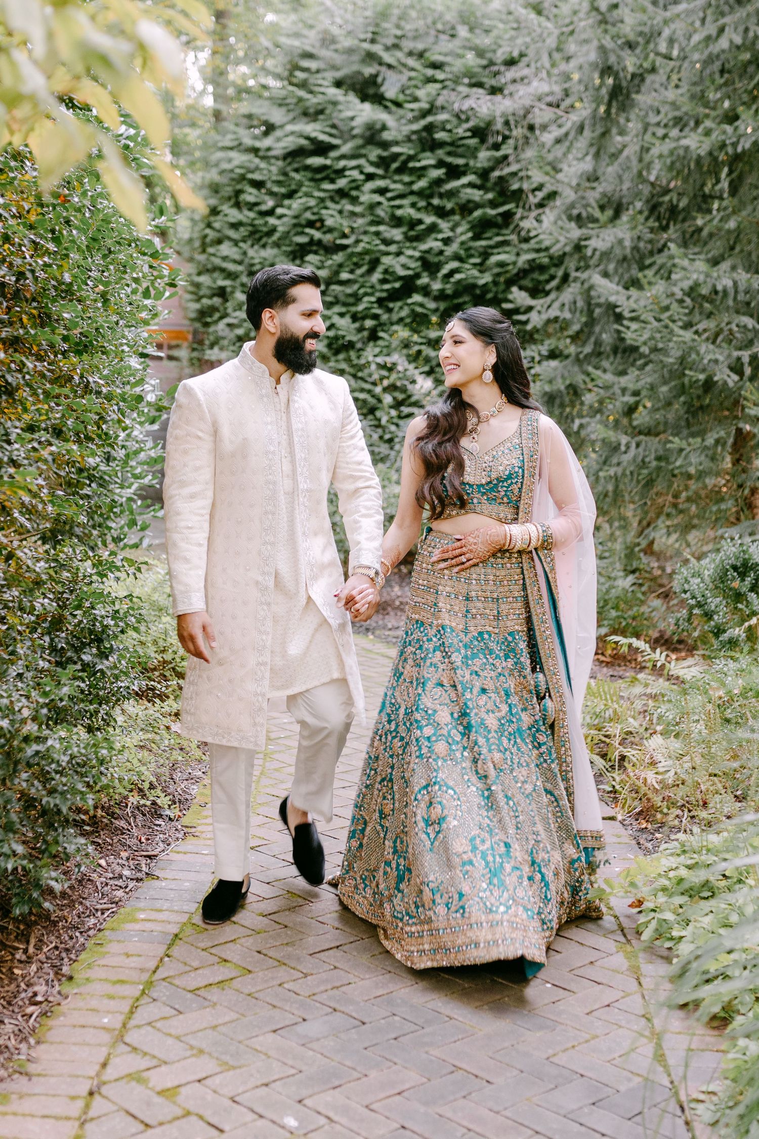 Couple in traditional Indian attire walking hand in hand on garden path surrounded by greenery.