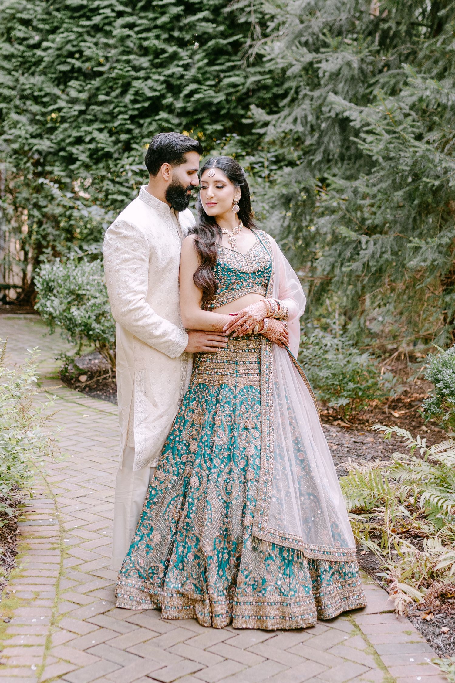 Romantic couple pose in traditional Indian wedding attire on brick pathway surrounded by evergreens.